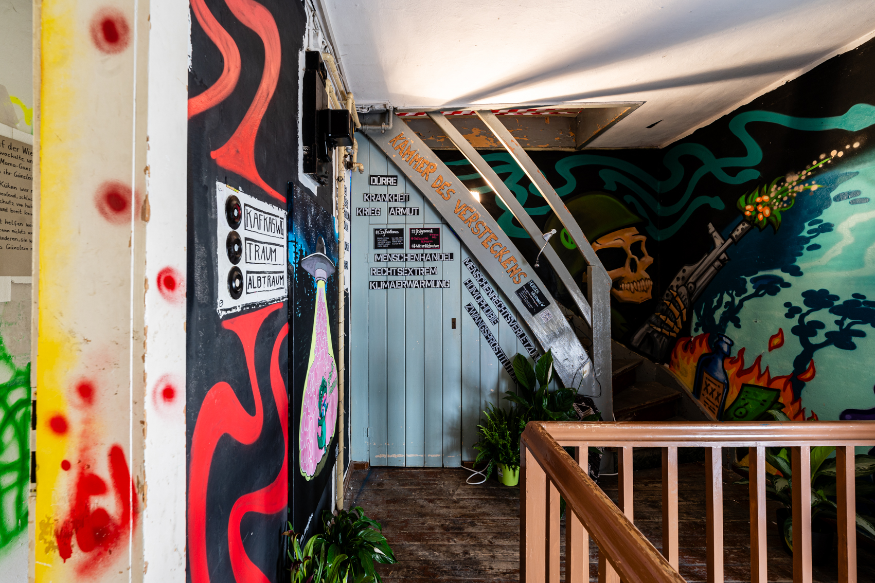 Bild 1 zu Kammer des Versteckens: Oberstes Stockwerk des Treppenhauses von Haus 1. Rechts und links an den W&auml;nden Graffiti von Lou Zeh. Geradeaus befindet sich die Treppe, die nach links oben in den Dachboden f&uuml;hrt. Darunter eine kleine Kammer. Auf dieser steht in schwarz-wei&szlig;er Schrift "Kammer des Versteckens".