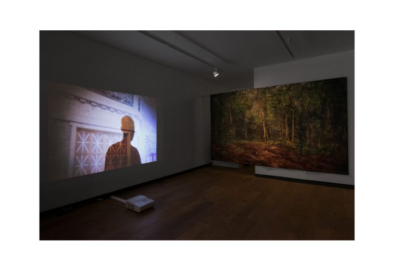 A photo of the gallery exhibition. The film "The Young Comrade" is projected on the left wall. A painting of a forest is hung on the back wall.