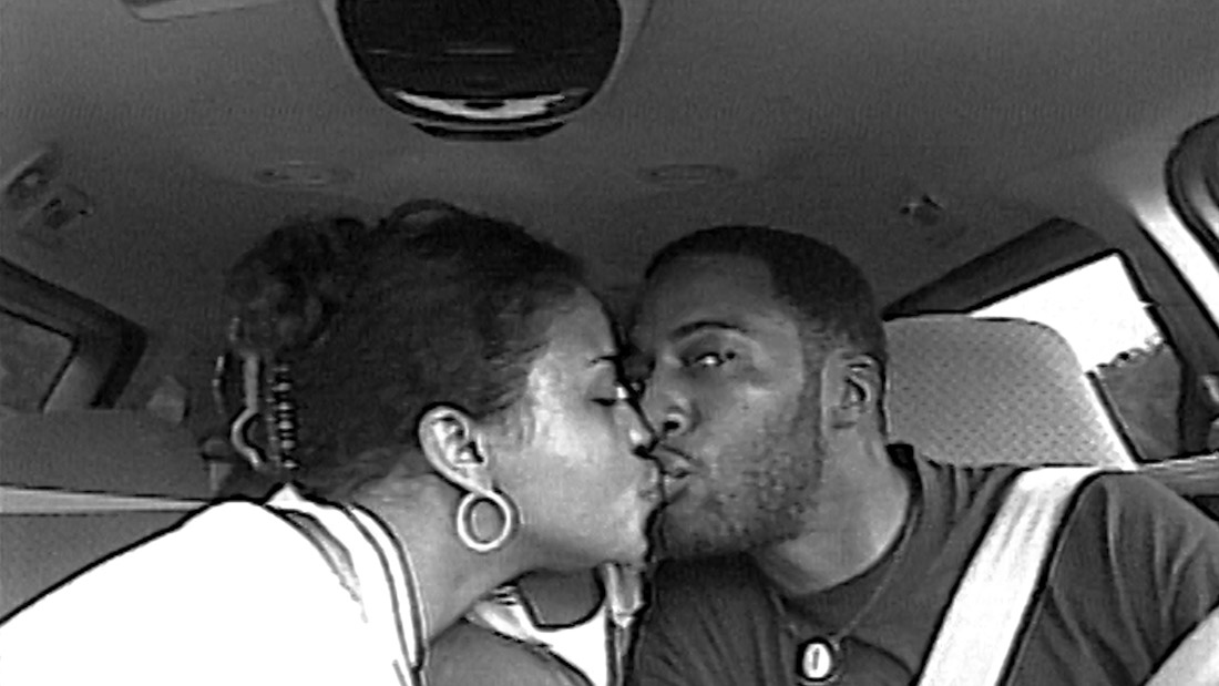 A grainy black and white photo shows a Black couple in the middle of kissing inside of a car, with the bearded person to the right (presumably driving) looking directly into the camera. The person on the left has their eyes closed amidst the kiss, wearing a circular earring.