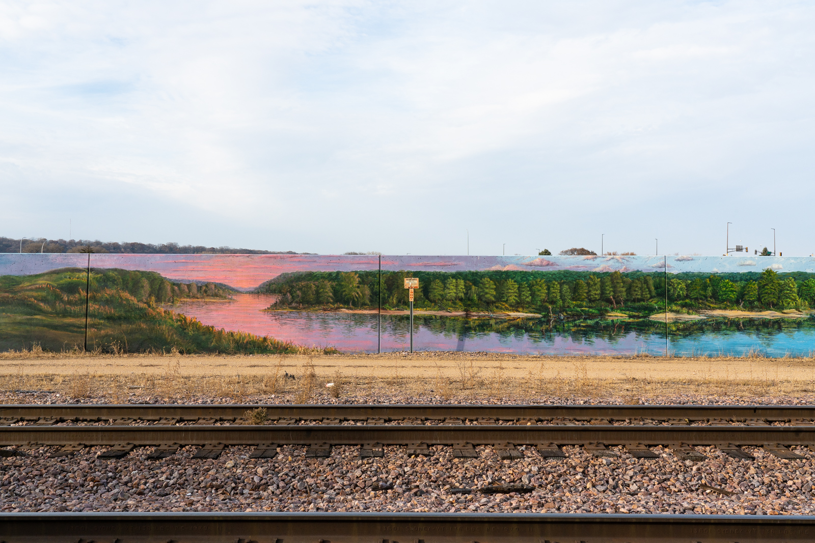 Along stretches of American farmland, which run parallel to the river, measures are taken to limit damage to critical infrastructure caused by catastrophic flooding, now common due to accelerated climate change. The Minnesota River, represented here as a mural, carries nitrogen and phosphate wastewater to the Mississippi River just beyond this flood wall in Mankato, Minnesota.