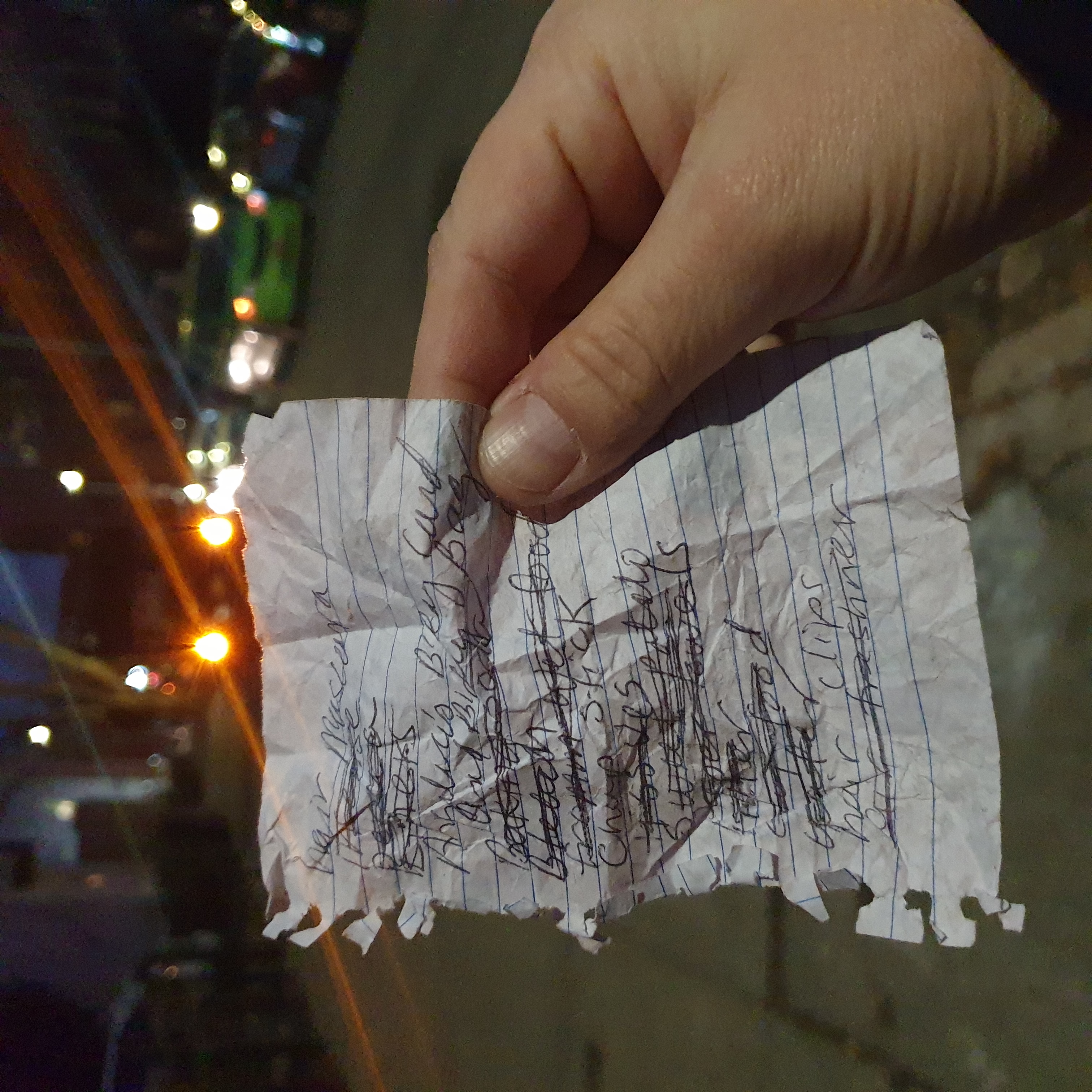 A hand holds up a crumpled piece of notebook paper with various things scribbled on it and crossed out. In the background car lights are visible on the street.