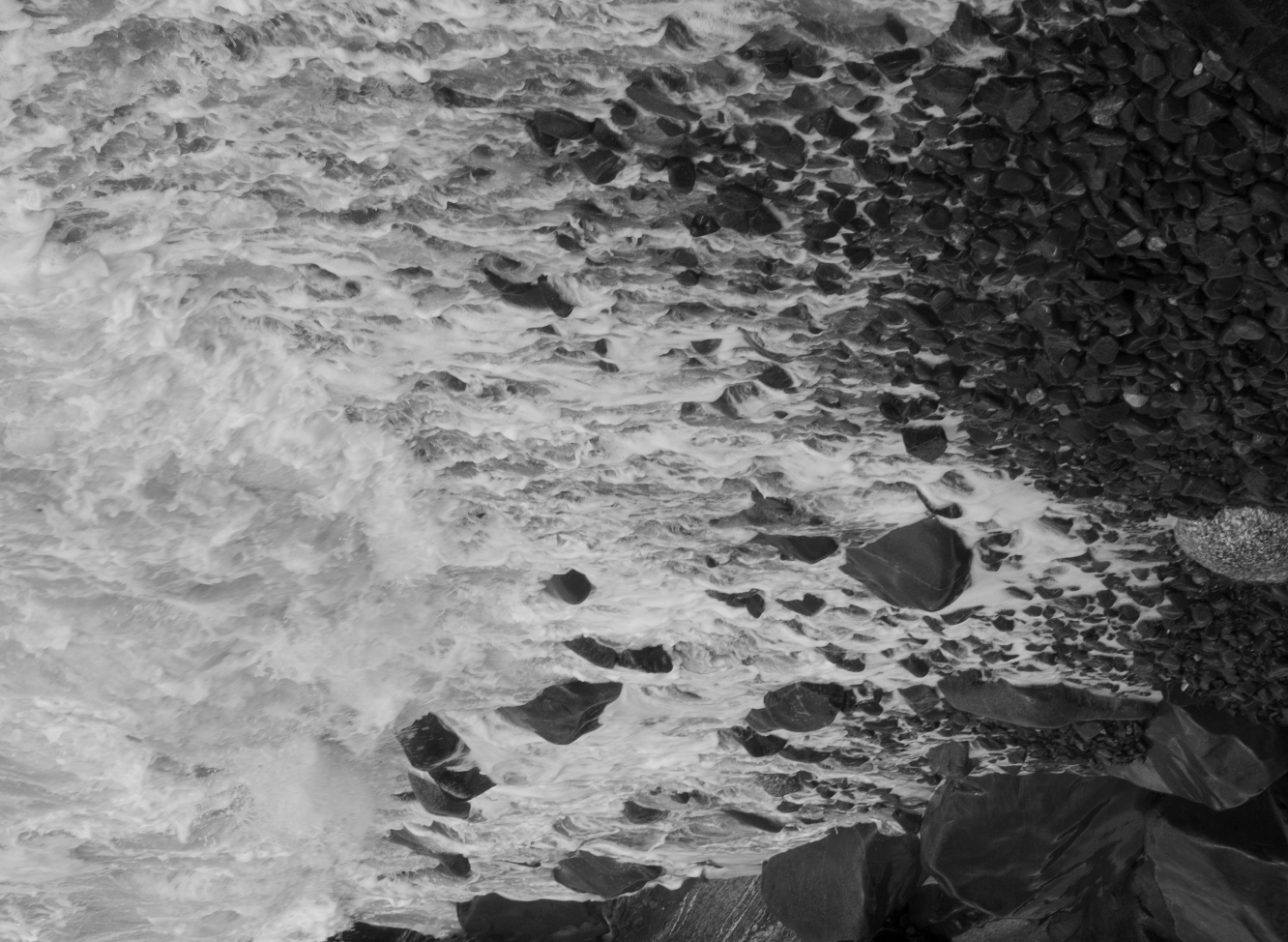Black/white photograph taken by Amy Schuessler: shown upside down, ocean tide crashing in against rocks on the shore