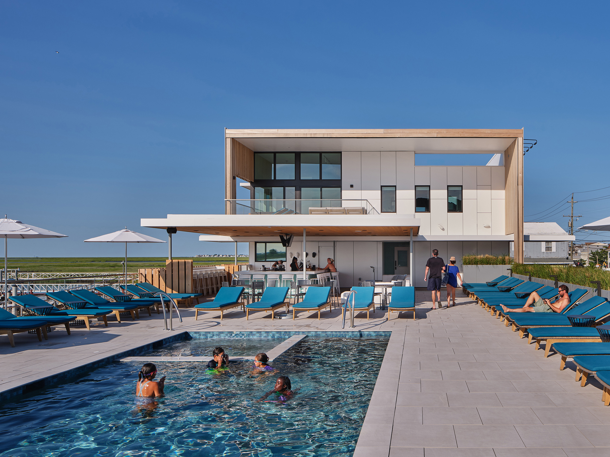 Exterior render of the Brigantine Paddle Club's elevated pool deck with several children swimming and members relaxing on blue lounge chairs next to the club building.