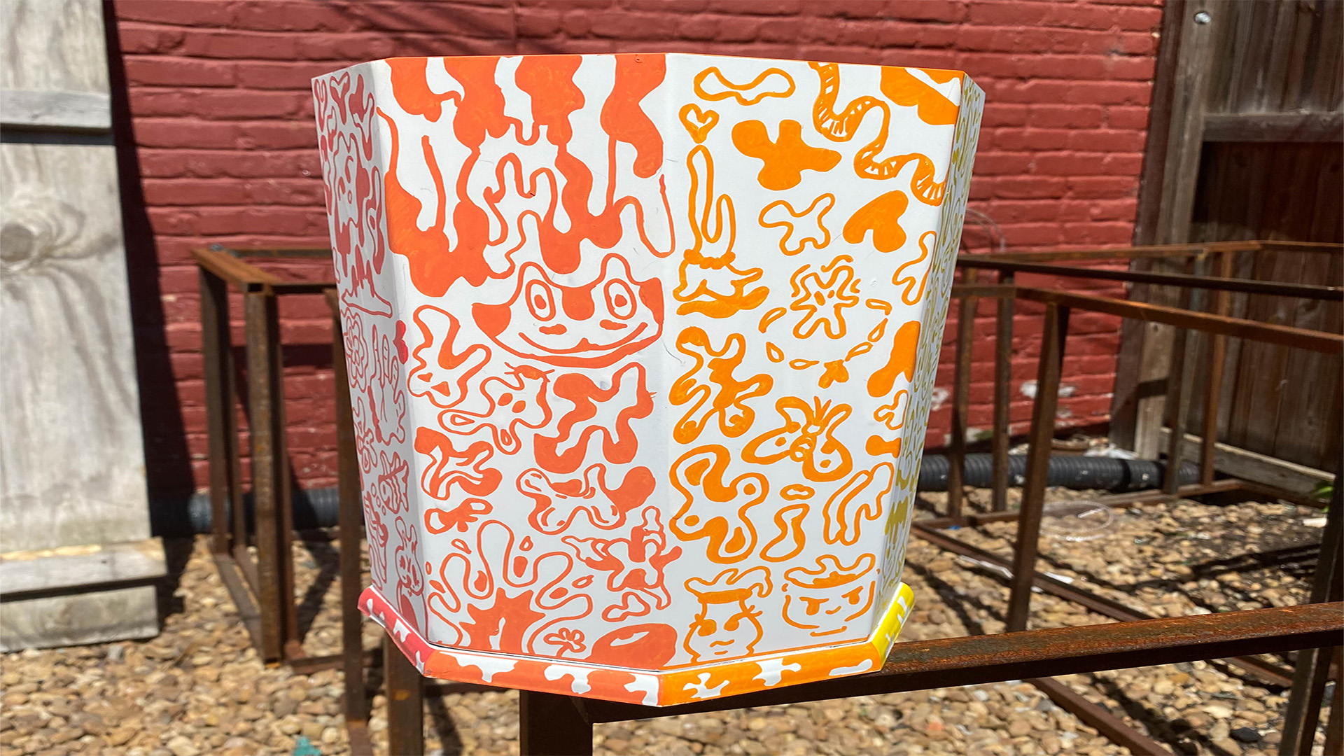 An image of an octagon trash bin outside on a sunny day. The bin has two sides covered in orange and light orange doodles. The doodles are made up of abstract shapes, faces, and objects.