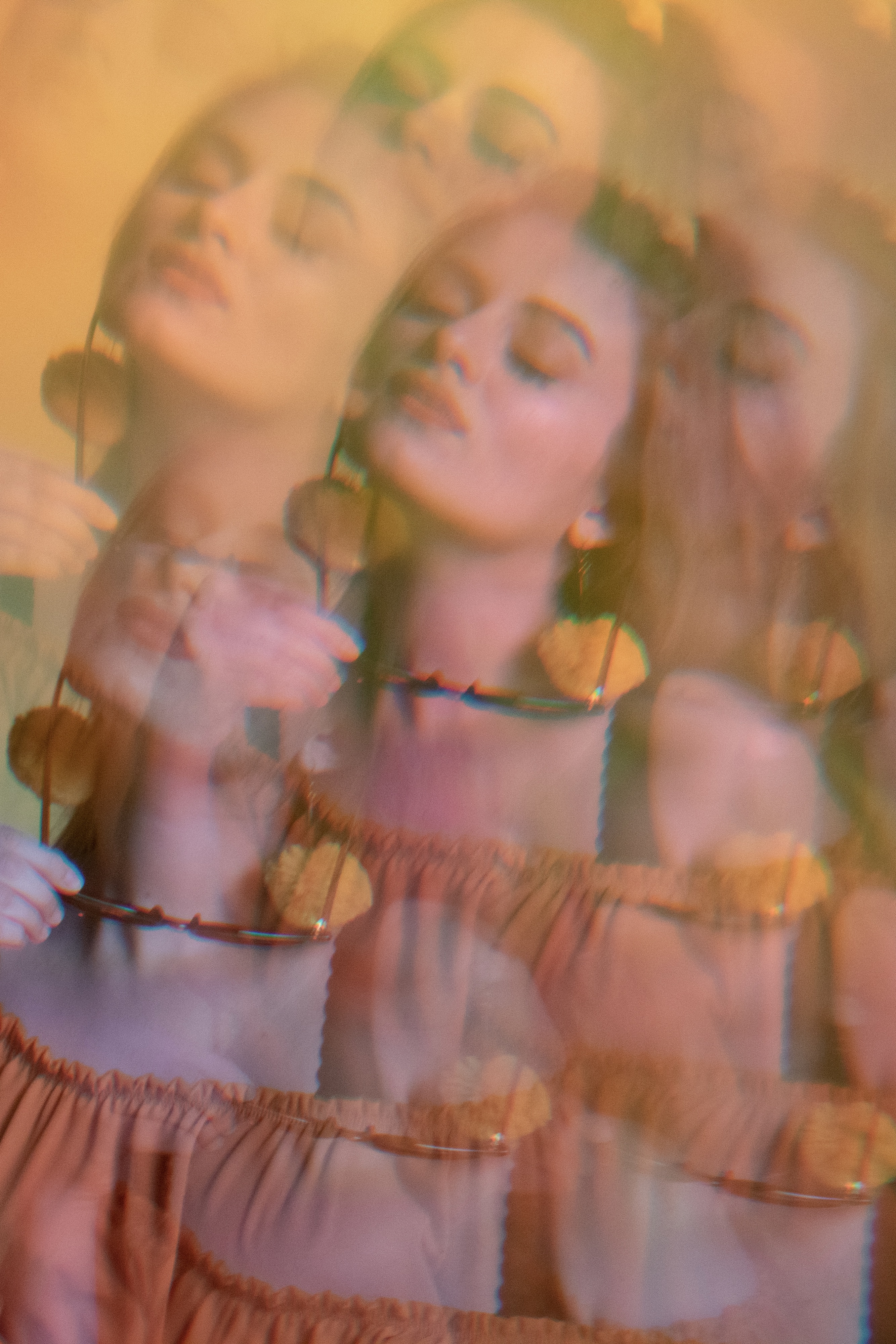 Dreamlike editorial image of a woman seen through a kaleidoscope, holding sunglasses in her hand with closed eyes and a serene expression. The composition is bathed in warm orange and yellow hues, enhancing its ethereal and surreal quality.