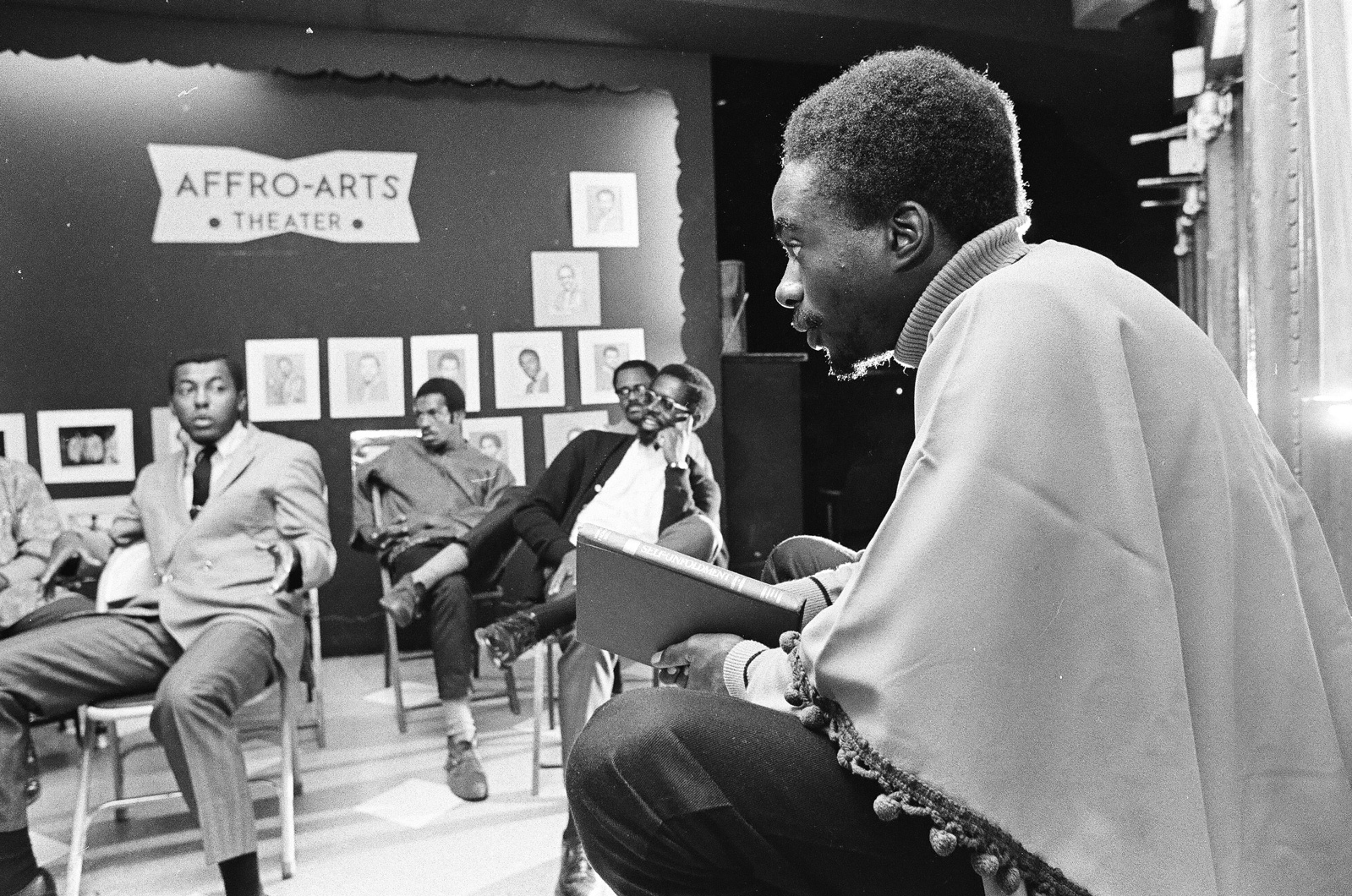Image: A meeting at the Afro-Arts Theater, 1968. The theater was founded by Kelan Philip Cohran in 1967 and was located at East 39th Street and South Drexel Boulevard, Chicago, IL. In the photo, a person sits close to the photographer, profile view, looking at others in the meeting, some who can be seen seated in front of a wall of portraits and a sign reading “Affro-Arts Theater.” Chicago Sun-Times Collection, Chicago History Museum. Photographer: Unknown.