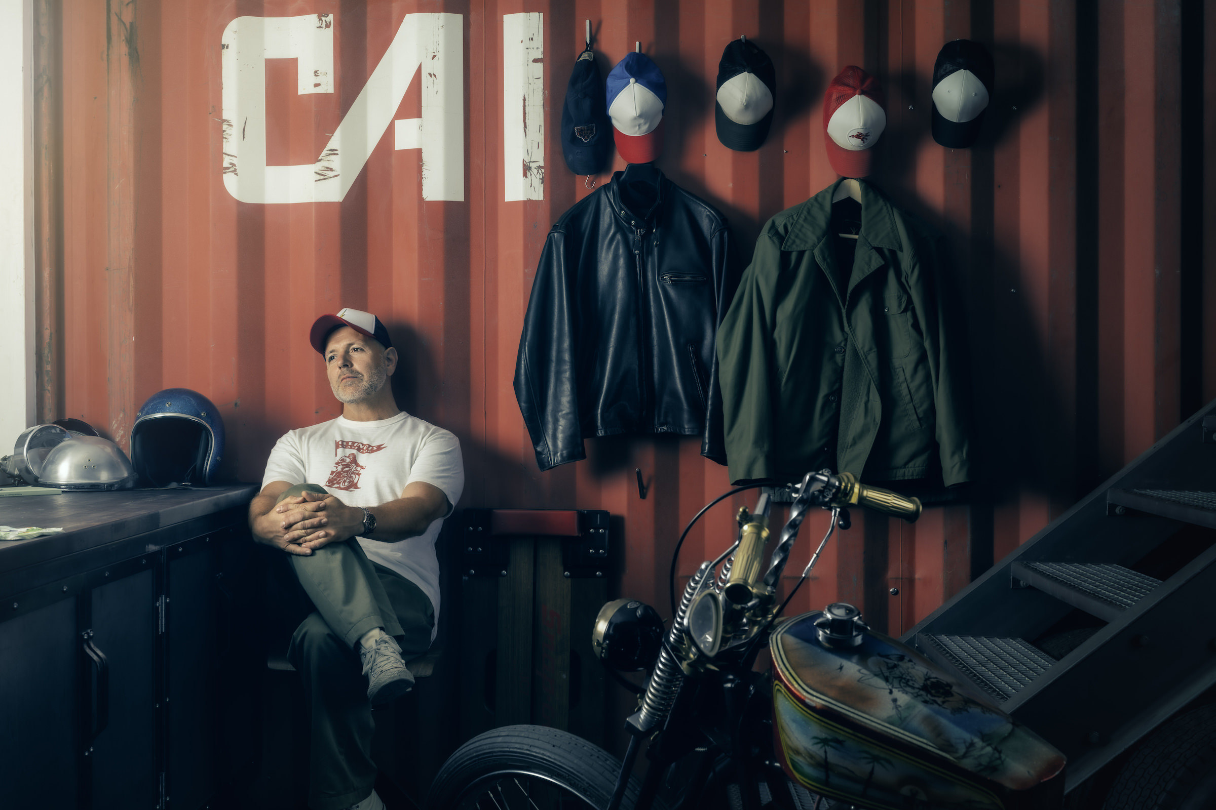 Environmental Portrait aus der Serie The Workshop. Stylische Werkstatt in Containerbauweise. Sitzend an einer roten Blech-Wand mit Aufschrift sieht man einen Mann, im Vordergrund ein 70er-Jahre Harley-Davidson Chopper.