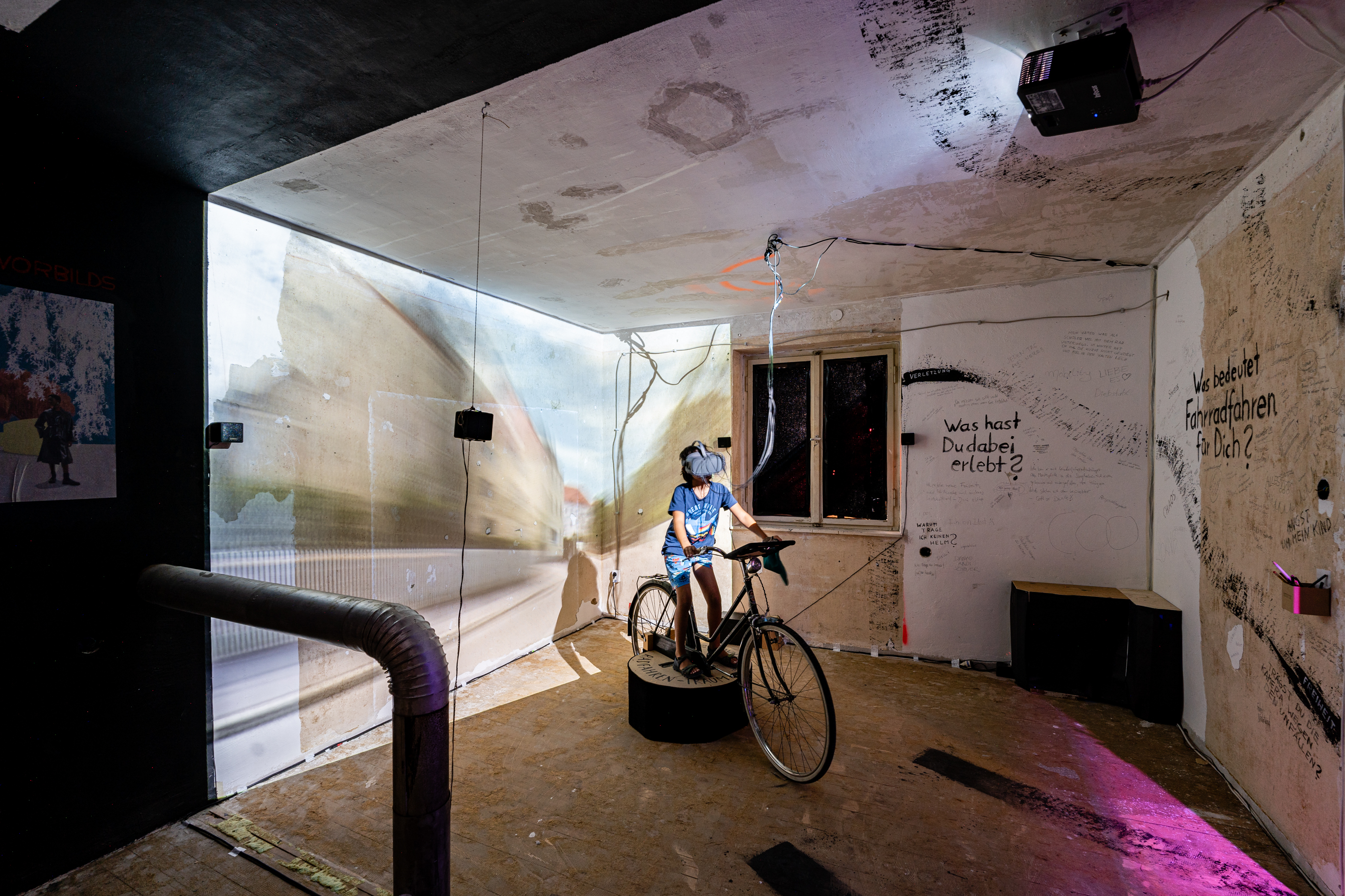 Bild 2 Radfahren:  Ein Kind sitzt mit einer Virtual Reality Brille auf dem Rad. An der rechten Wand k&ouml;nnen Besucher:innen festhalten, was sie beim Radfahren erlebt haben und was Fahrradfahren f&uuml;r sie bedeutet.