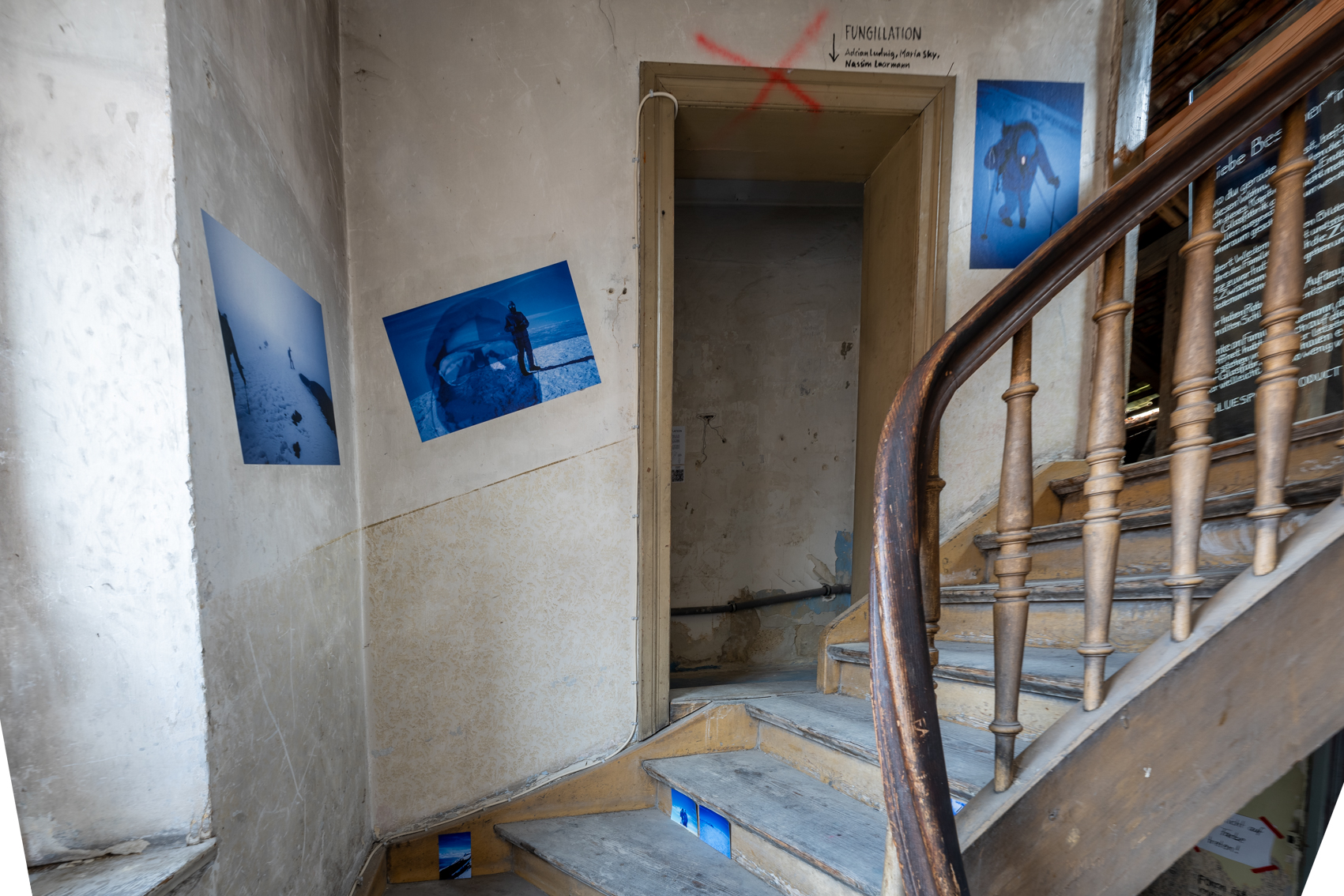 Bild 2 zu Stairway to heaven: Links des Treppenaufgangs an der Wand h&auml;ngen weitere Fotografien in etwa DinA2 Gr&ouml;&szlig;e. Mittig der Wand befindet sich eine T&uuml;r, die in einen kleinen Raum f&uuml;hrt, von dem aus die Installation Fungillation durch ein Guckloch in der Wand sichtbar ist. 