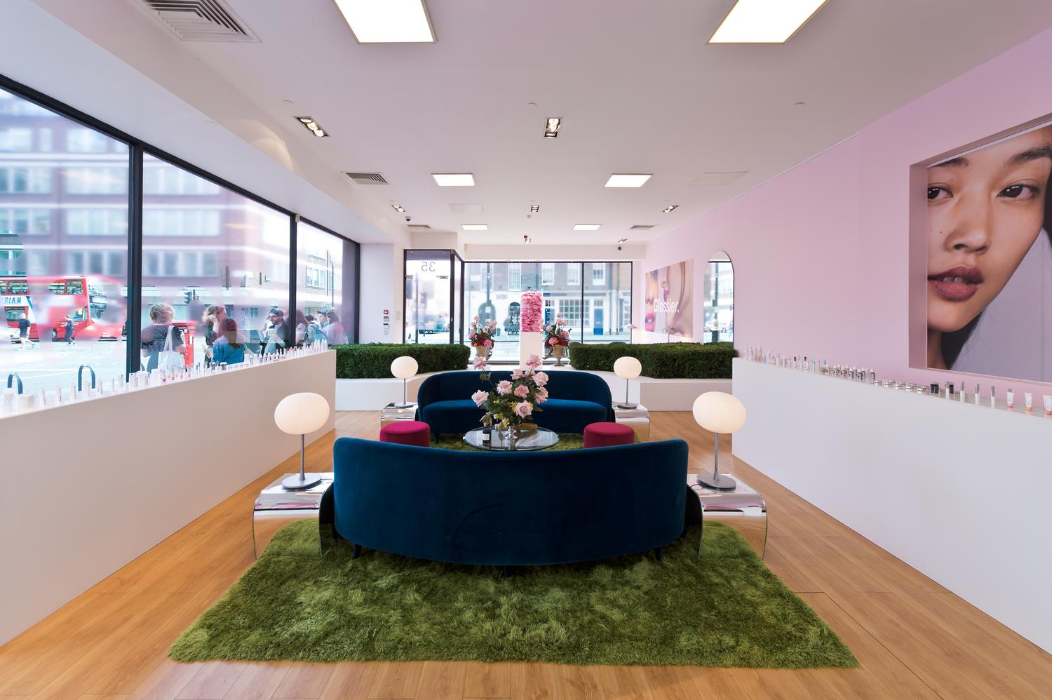 Glossier London pop-up interior with blue sofas, pink walls, large model portraits, and product displays.