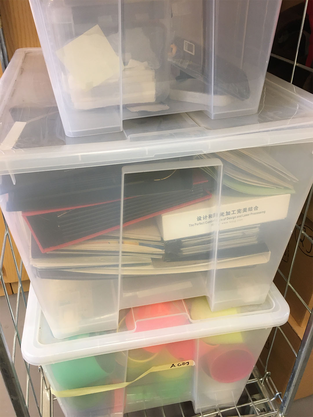 Three partly see-through plastic boxes are stacked on top of each other. They include various items, such as a phone, folders, papers, and brightly coloured unidentified objects.