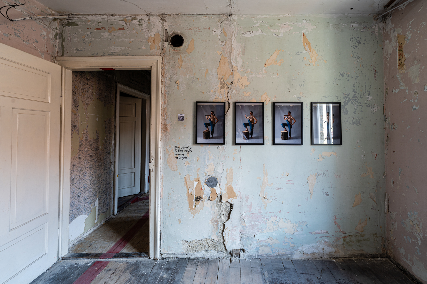 Bild 3 zu The Beauty and the Boys: Blick auf die Wand rechts der T&uuml;r mit abbl&auml;tternder Wandfarbe. Vier gerahmte Fotografien in einer Reihe zeigen eine m&auml;nnlich gelesene Person mit Latzhose. Das rechte Bein steht auf einem Holzpflock. Der rechte Arm ist angewinkelt und h&auml;lt jeweils einen anderen Gegenstand wie zum Beispiel eine Axt oder einen Holzpflock auf den Schultern. Der linke Arm ist in die H&uuml;fte gest&uuml;tzt.