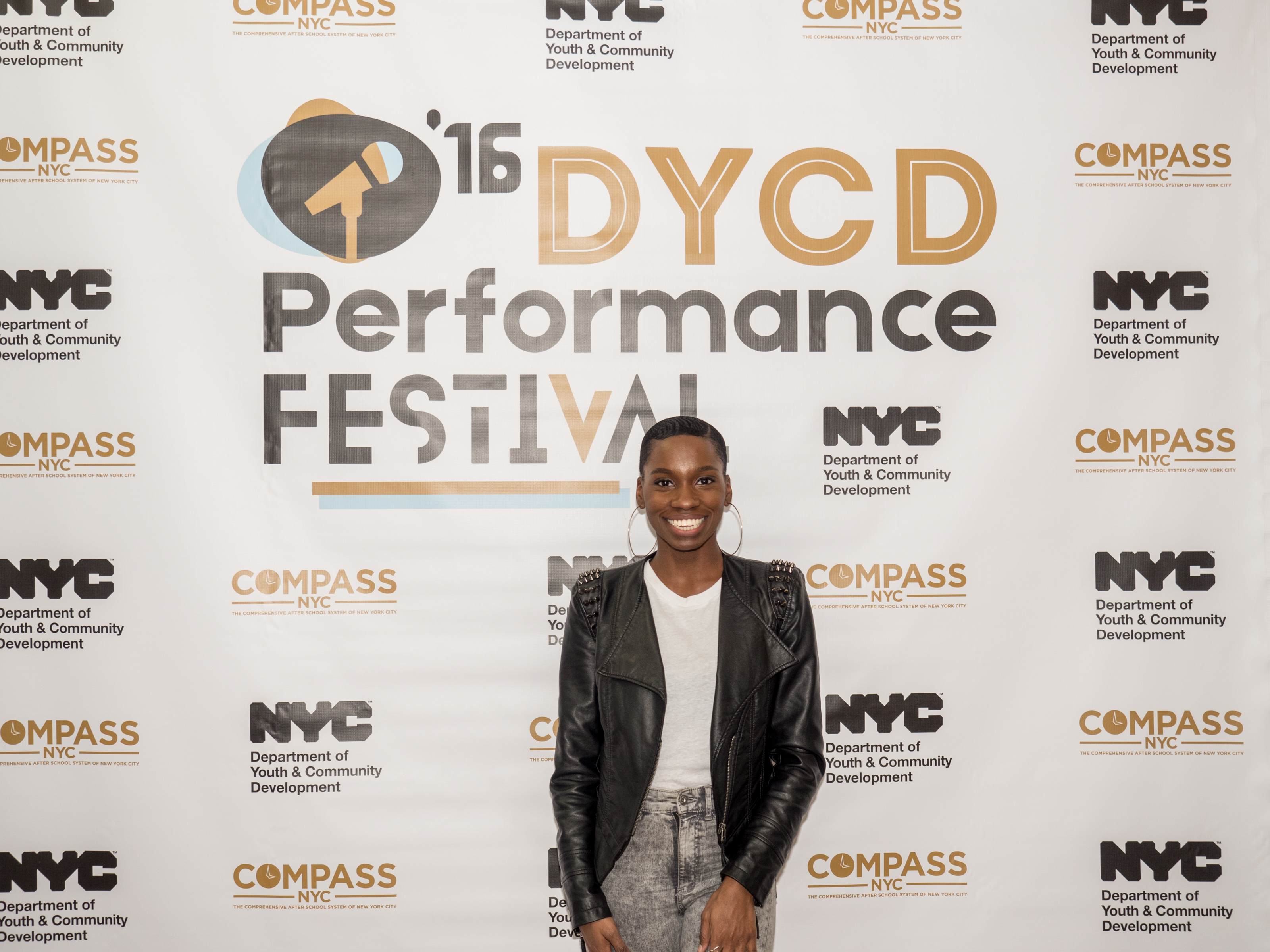 A guest posing in front of a white step and repeat with the DYCD Performance Festival logo large and centered, the DYCD and COMPASS logos repeat in a pattern around the Performance Festival logo.