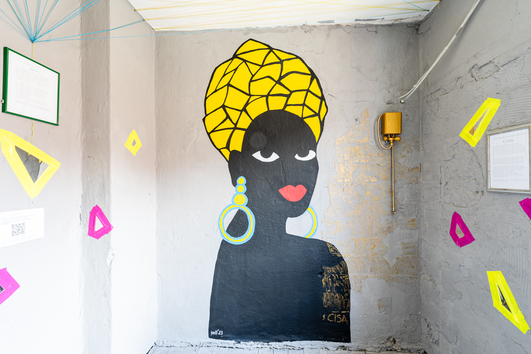Bild 1 zu Ein Schritt zur&uuml;ck nach vorn: Gro&szlig;e Wandmalerei auf hellgrauer Wand. Oberk&ouml;rper einer Person mit gelber, aus geometrischen Formen zusammengesetzter Kopfbedeckung und gro&szlig;en gelben Ohrringen. Sie Blickt &uuml;ber die Schultern den Betrachtenden an.