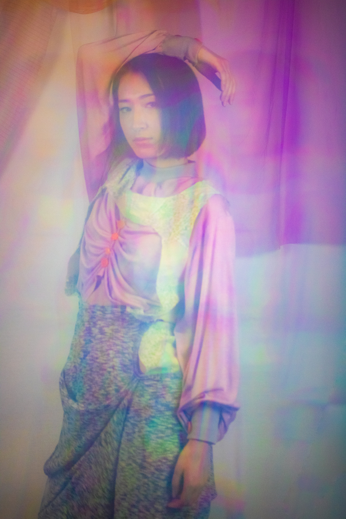 Three-quarter shot of a woman in a grey skirt, purple top, and green vest, standing with her hands behind her head. She is positioned in front of a colorful fashion collection, with a rainbow of light reflecting through the image, adding an ethereal, dynamic quality.