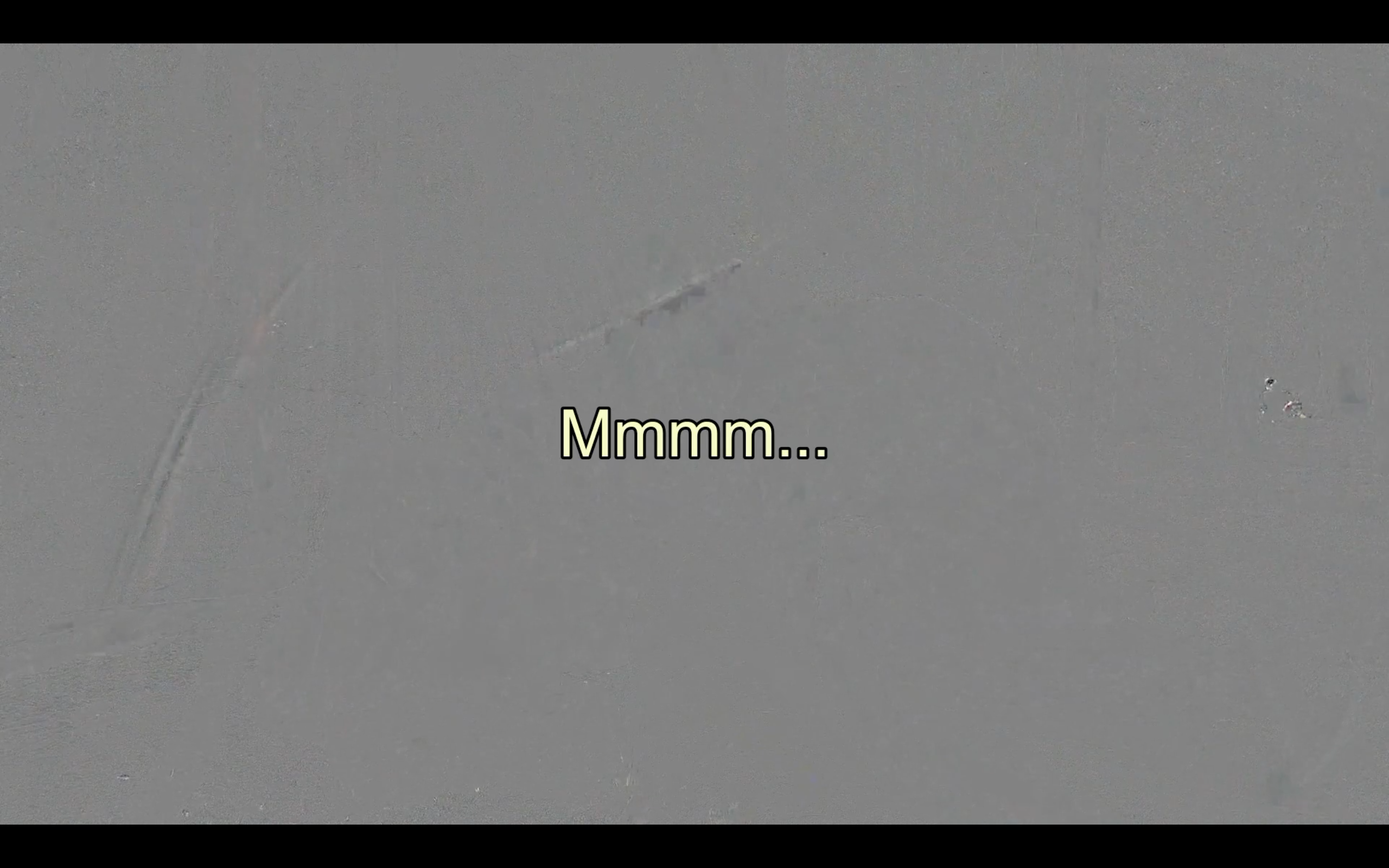 A video still. Light yellow text sits in the centre against a grey background. It reads, “Mmmm…”