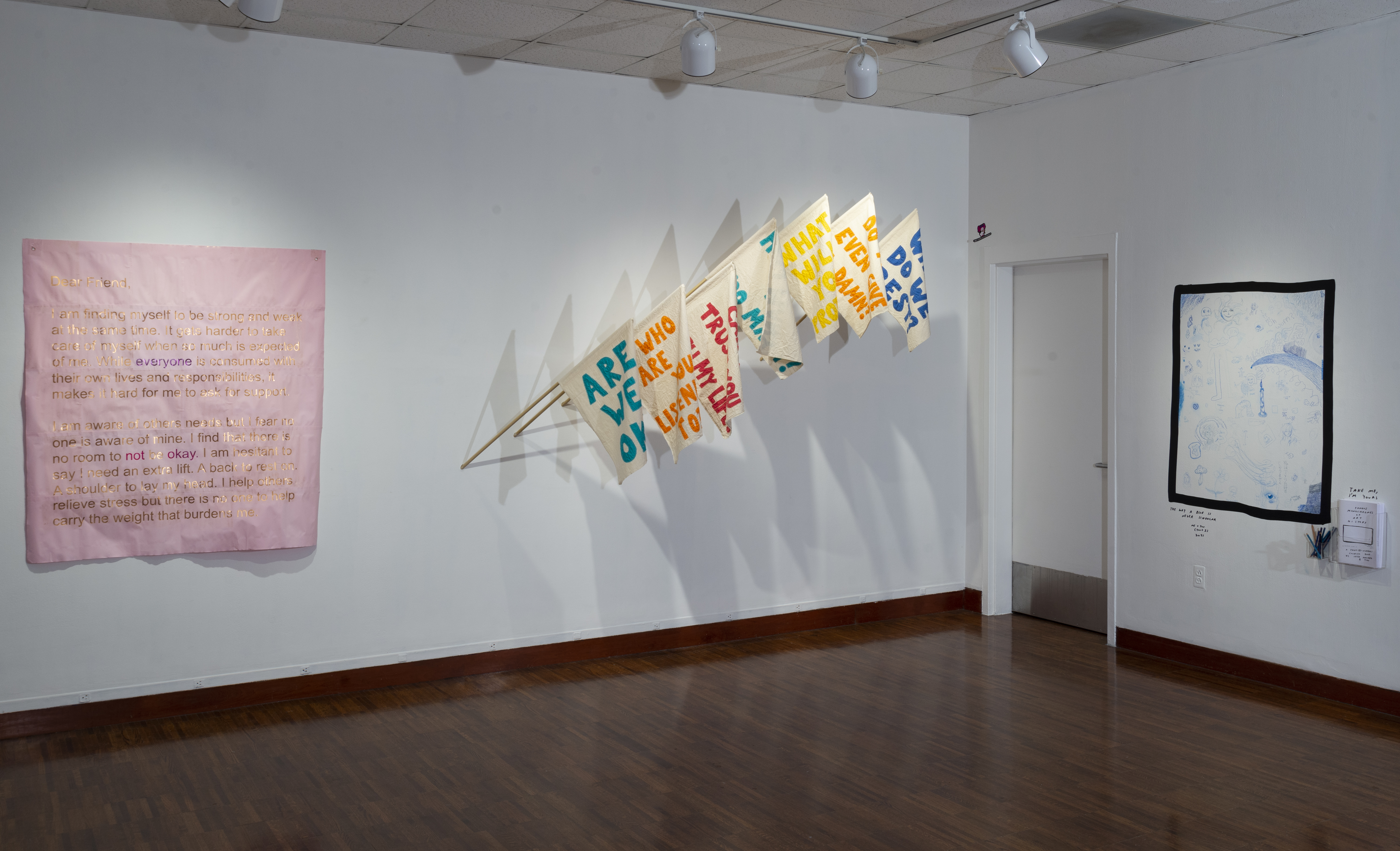 A corner of a white walled gallery with three pieces of art in frame. On the left a large, pink square of fabric that has illegible gold text. In the middle, an arc of 7 flag-banners that all read different messages in colored text on a canvas colored fabric. On the right less visible square of white with faint blue markings surrounded by a thick black border.