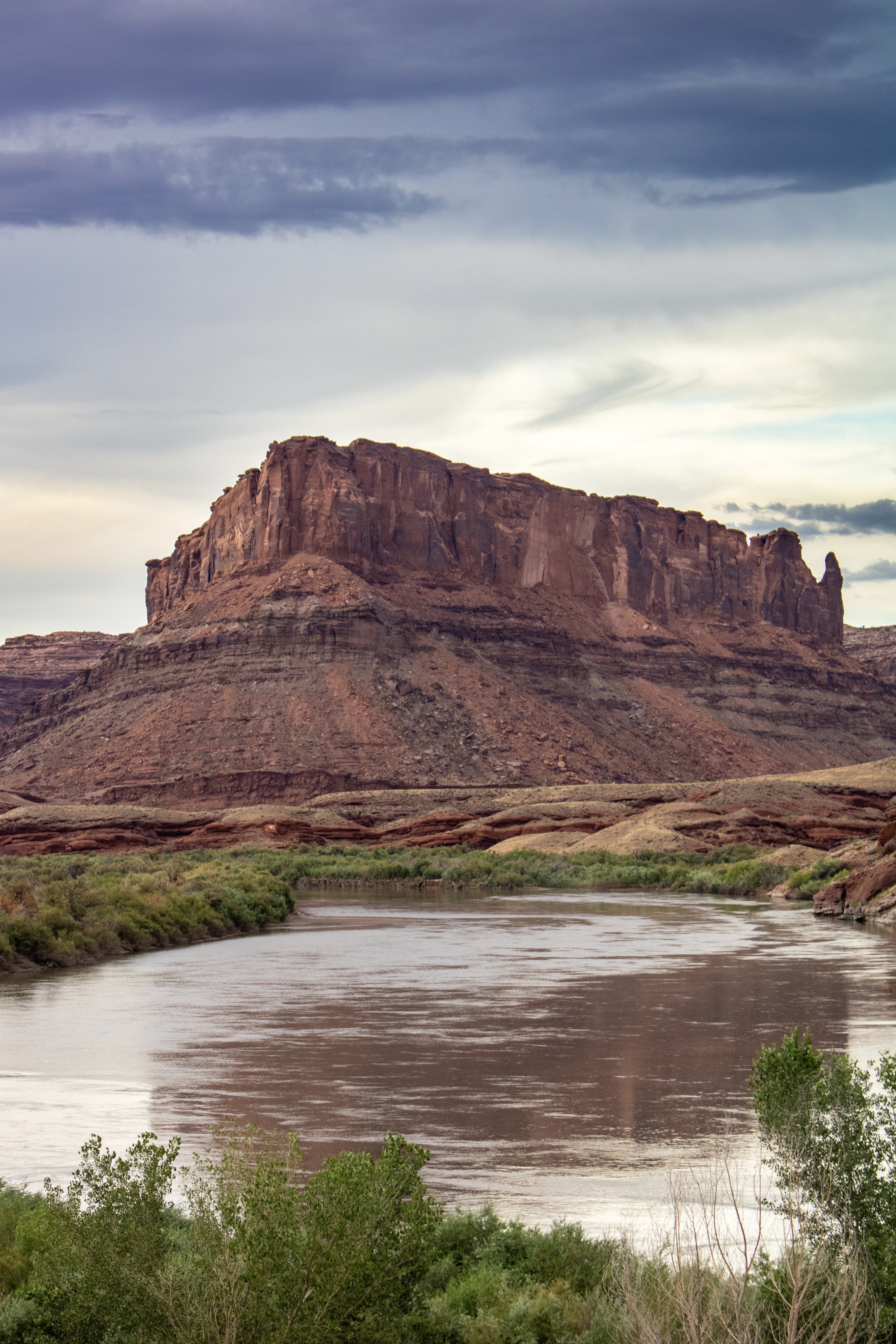 Dramatic sandstone butte under a cloudy sky with a winding river in the foreground, flanked by lush greenery and rocky terrain, conveying tranquility with the Colorado River at the Moab’s Colorado Riverway.