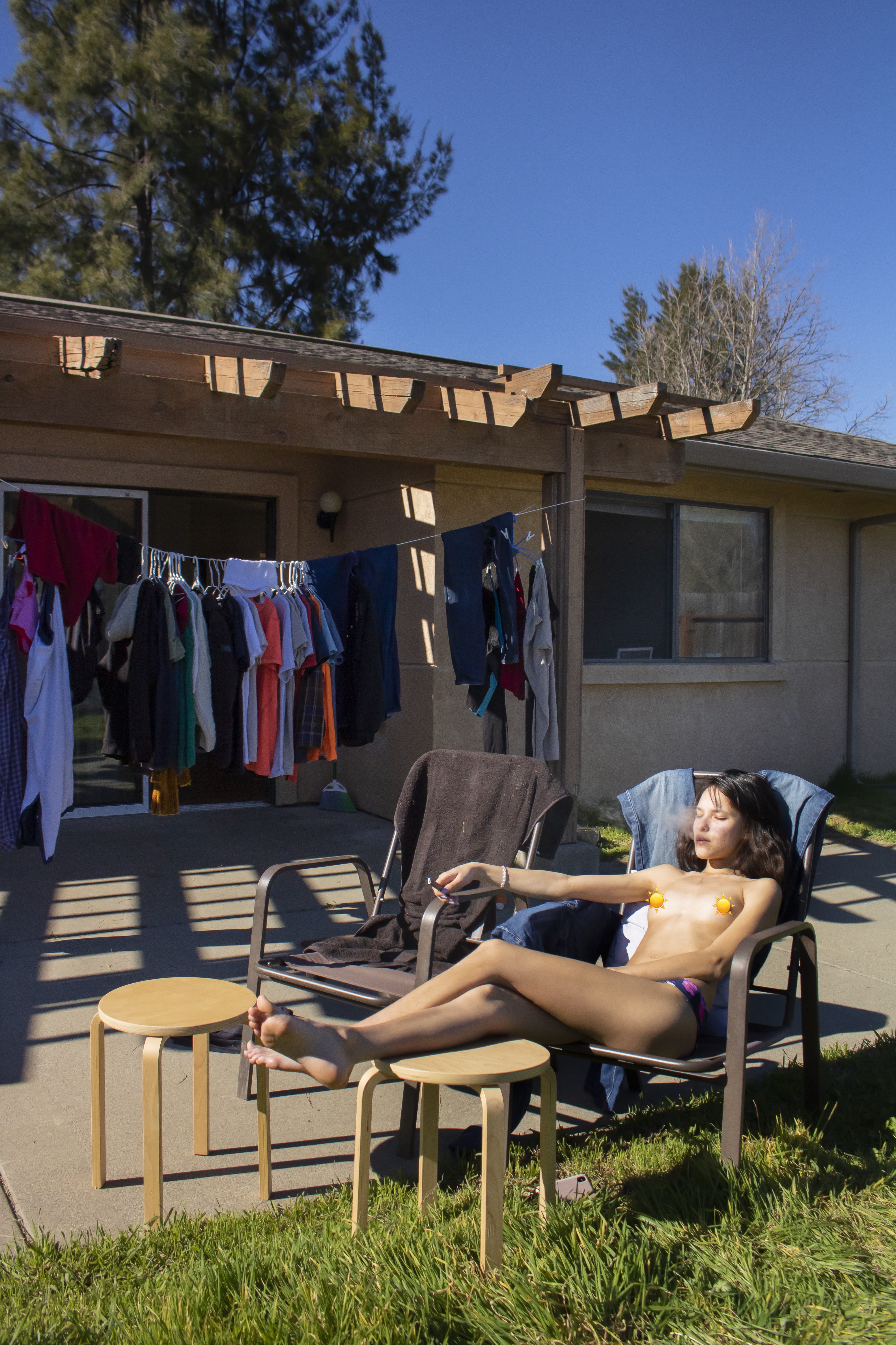Sun-drenched portrait of a topless woman sunbathing in her backyard, cigarette in hand, with clothes hanging on a clothesline behind her. A raw and intimate moment captured in golden light.