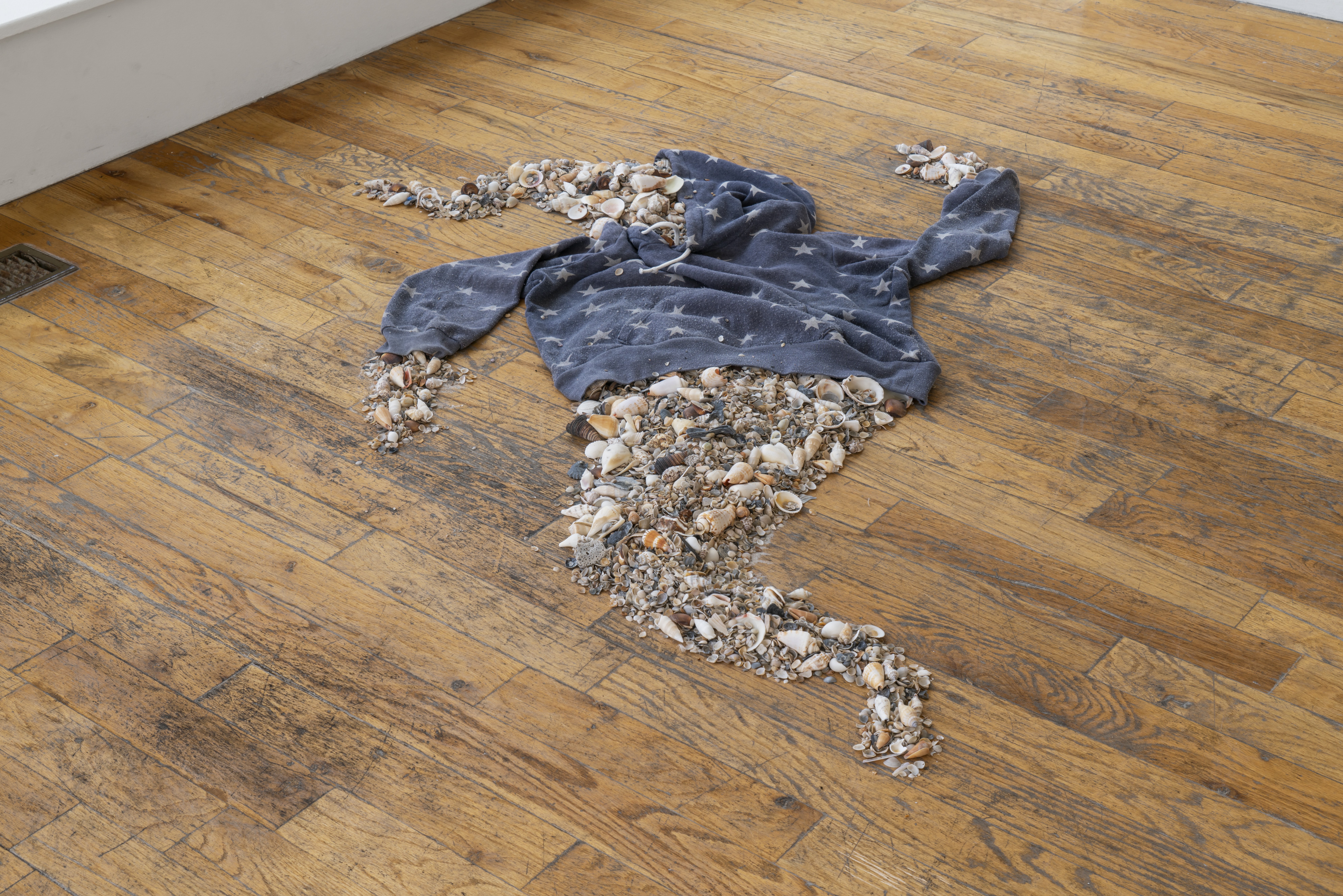 An empty hoodie with a faded star print pattern is lying on the ground, and coming out of the arms and torso it is a neatly organized pile of seashells, shaped to seems like a ghostly seashell figure is wearing the hoodie.