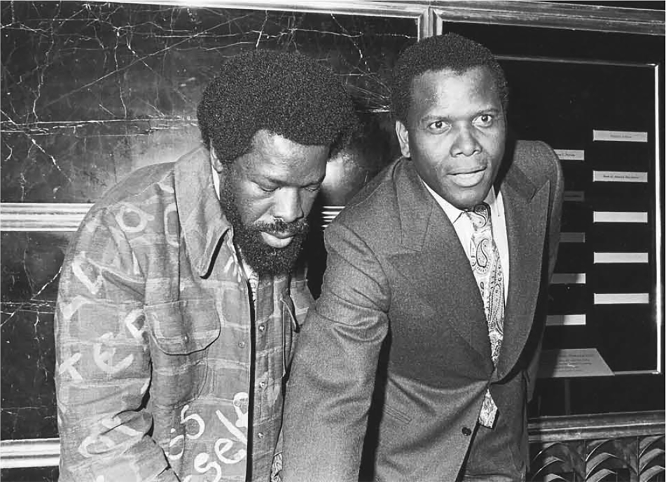 Casper Banjo helps Sidney Poitier make his handprints for the Black Filmmakers Hall of Fame, Inc., c. 1980. Banjo is wearing his signature jacket, printed with bricks and with the words “express yourself” visible on the front.