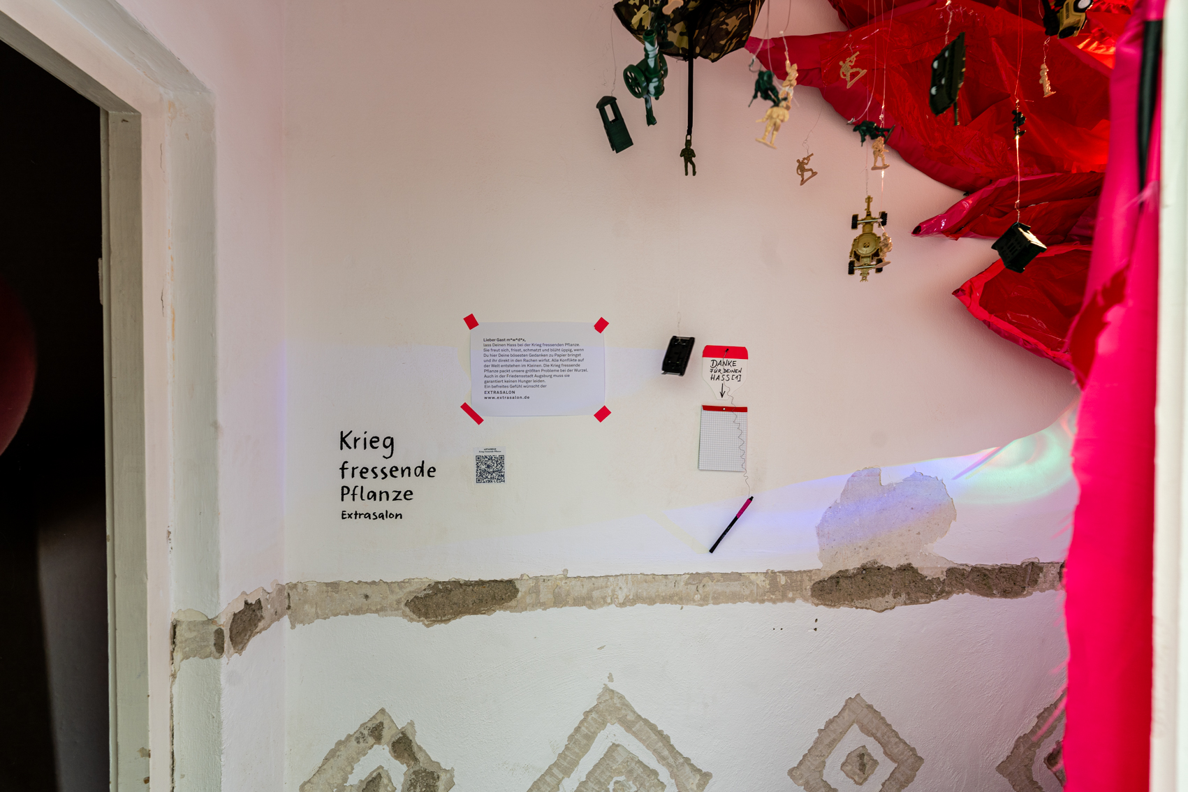 Bild 2 Krieg fressende Pflanze: Blick im Raum stehend auf die linke Wand, auf der Titel und K&uuml;nstler:innen Name steht, sowie ein kleiner Infotext. Ein Block mit der &Uuml;berschrift: "Danke f&uuml;r Deinen Hass". In der rechten oberen Bildh&auml;lfte sieht man deutlicher die kleinen Plastikfiguren, die von der Decke h&auml;ngen, sowie die roten Stoffbl&auml;tter der Pflanze.