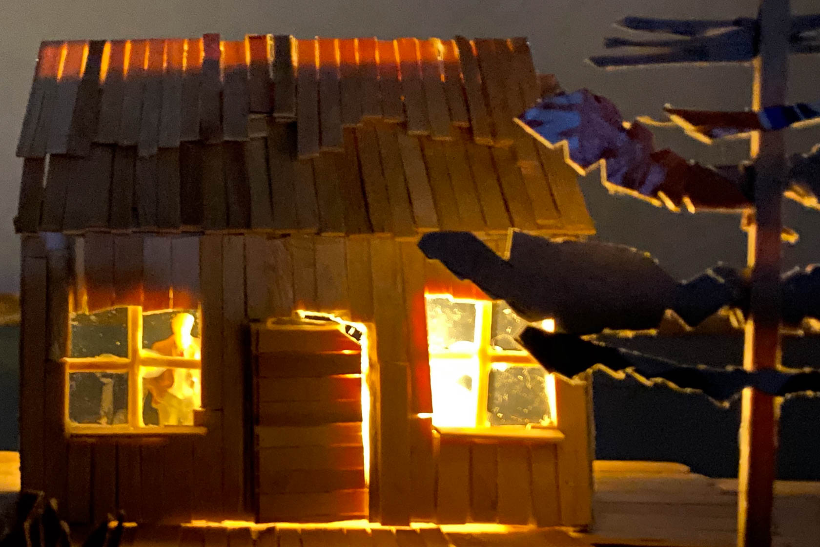 A miniature home lit from inside with a full moon above with whimsical, fairy-tale feel.