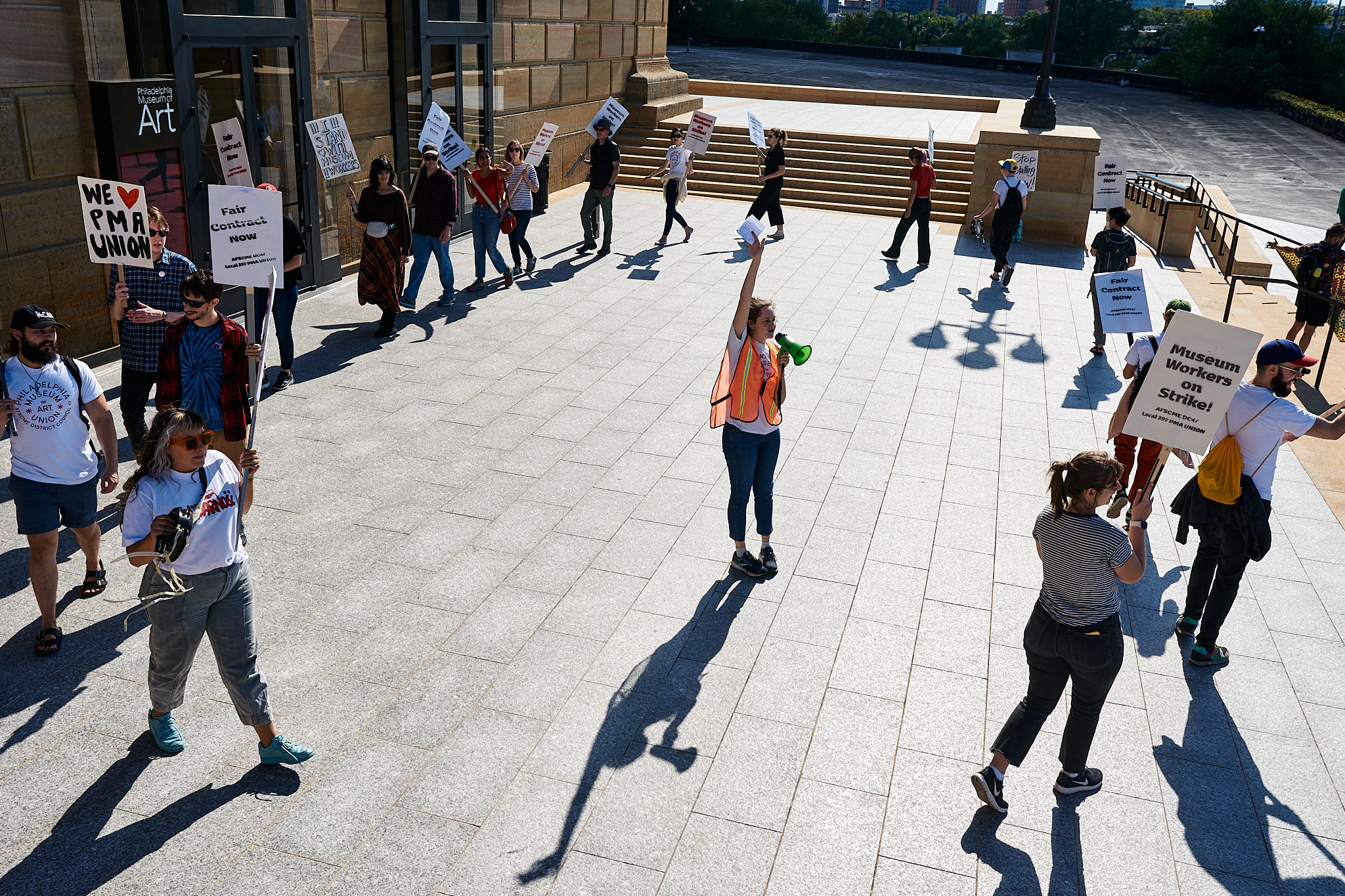 Color photo of a group of 18 striking workers holding picket signs in front of an entrance to the Philadelphia Museum of Art, and moving in a circle around a single worker in an orange safety vest and a small green megaphone mid-sentence, seen from slightly above.