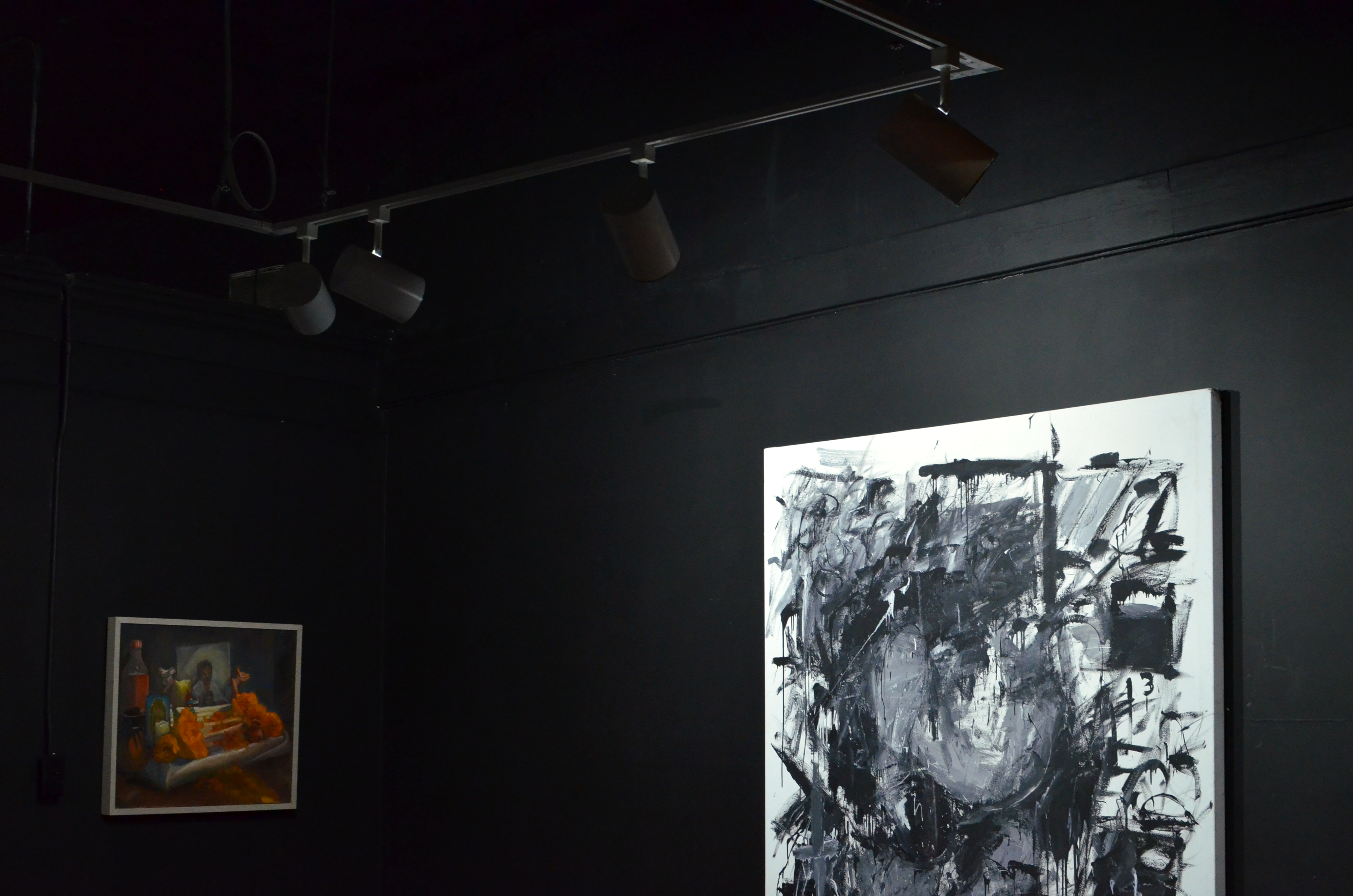 A color photograph of a corner in a dark gallery room with painted black walls and a ceiling lit with several track lights. On either side of the corner are a pair of paintings, one smaller colorful on the left, and one larger and monochrome on the right.