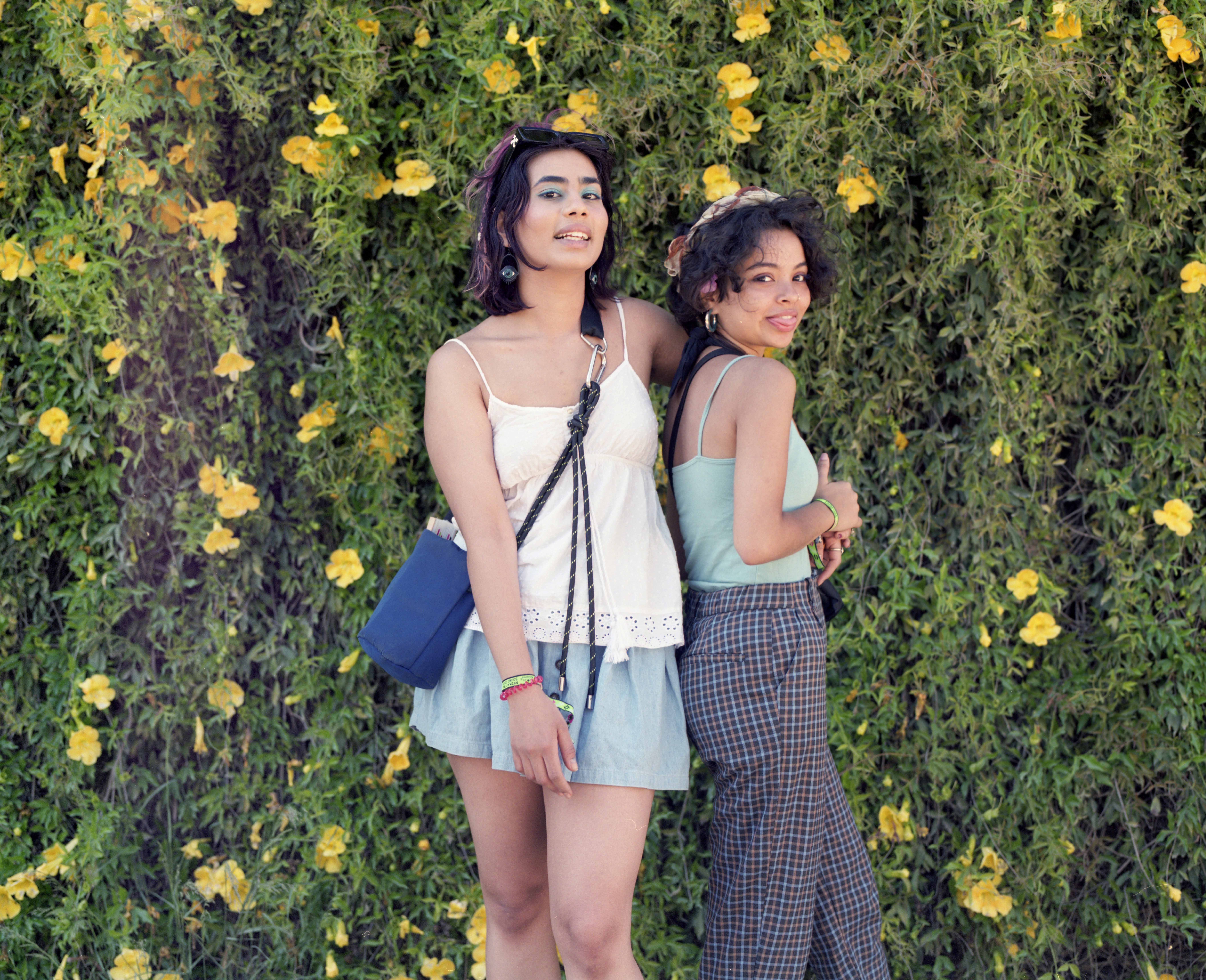 Full body portrait of two girls, stood in front of green hedge with yellow flowers. One wears checkered pants and blue top and the other wears blue skirt and white top