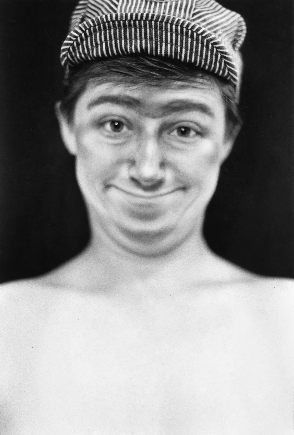 A photorealistic greyscale image of a shirtless figure with a light skin tone, short dark hair, and pinstripe hat, seen from the chest up, looking directly forward and has a smiley, bemused look.