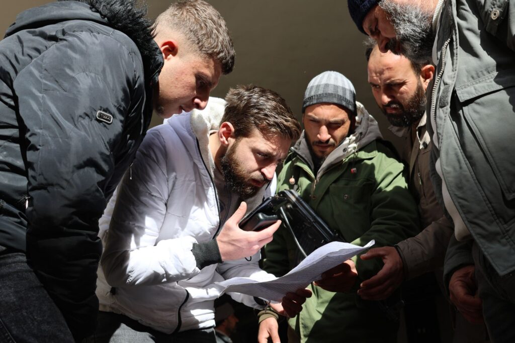 A group of men take photographs of papers they found on the ground, hoping to find the names of their missing loved ones.
