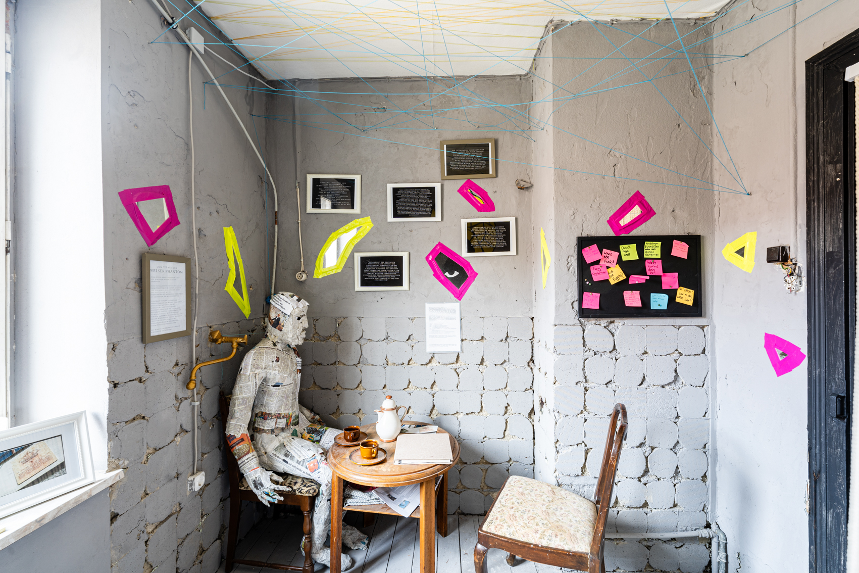 Bild 4 zu ein Schritt zur&uuml;ck nach vorn: Blick an Wand rechts im Raum. Eine Lebensgro&szlig;e Figur aus Zeitungen sitzt an einem gedeckten runden Cafe Tisch. Gegen&uuml;ber von ihr ein Leerer Stuhl. An der Wand dahinter h&auml;ngen einige Bilderrahmen mit Texten darin.