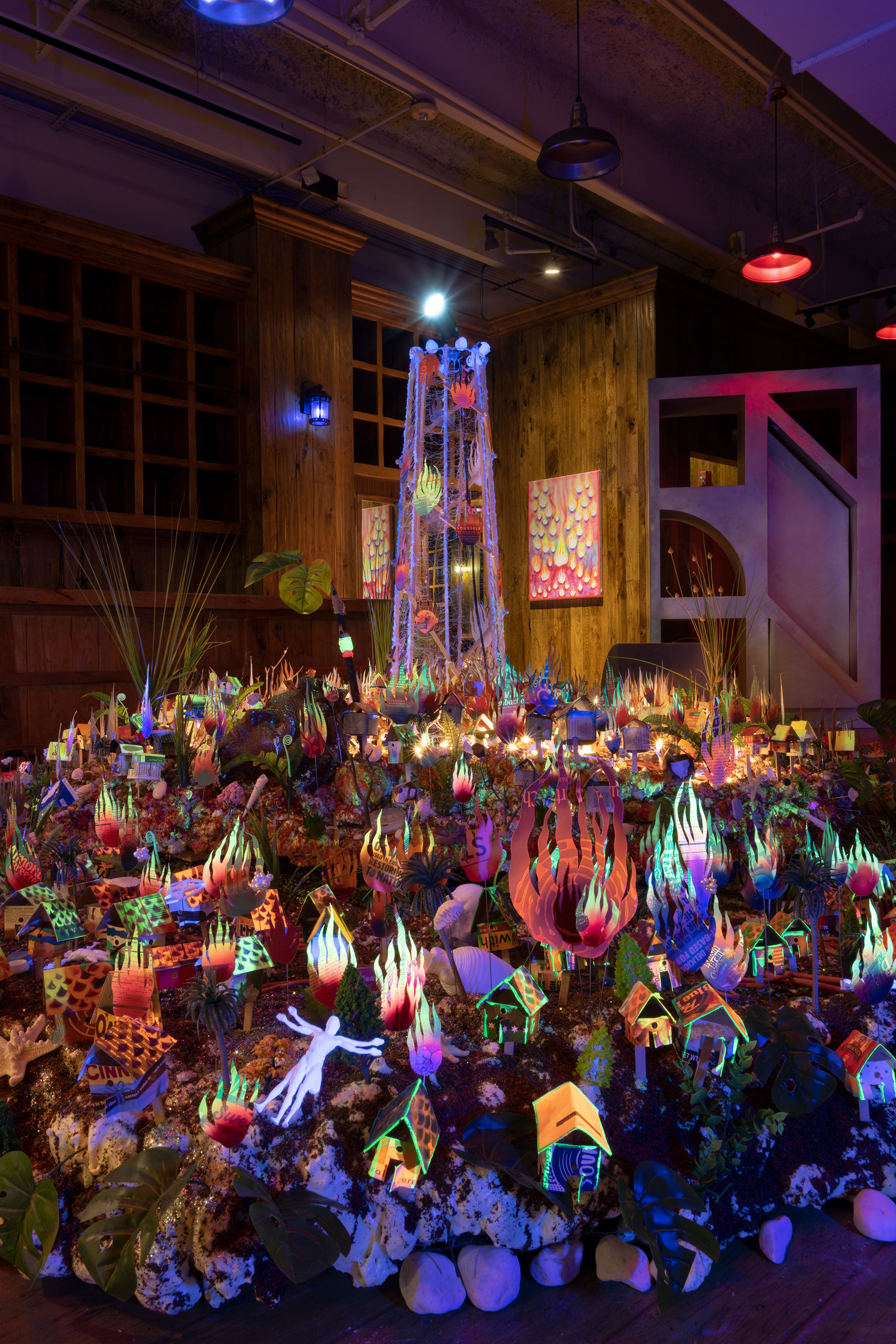 Orlando Estrada, The Infinite Vision in the Ethereal Rainbows Swarming Behind my Eyelids Reveals a Beacon, exhibition view, Bas Fisher Invitational and Bridge Initiative, Miami Beach, FL, USA. Tiered Panorama, a large multi-level sculpture stands in the center: a white tower peering from a dystopian mound under blacklight, Fire Painting #1 is displayed in the distance.