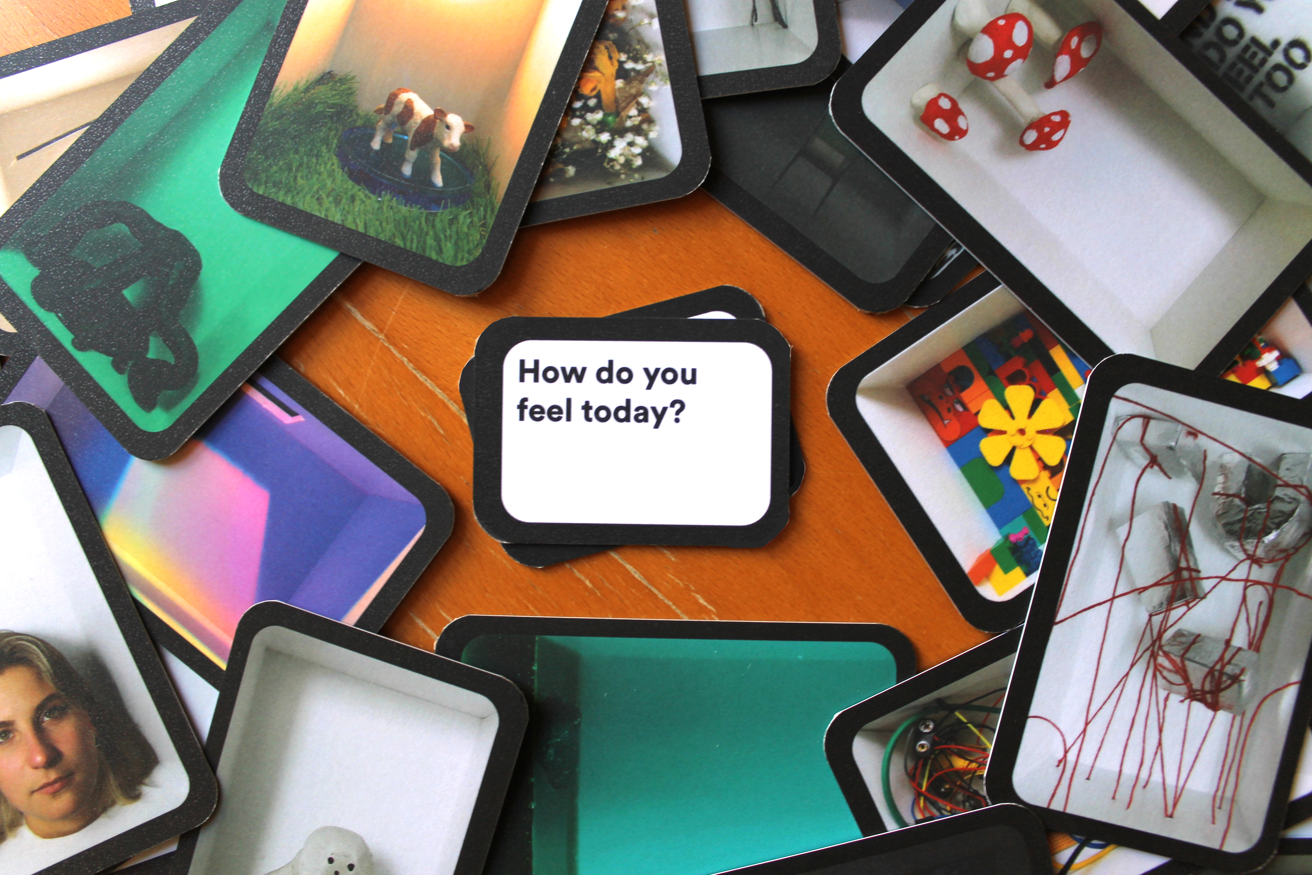 A set of game cards each depicting a different art installation are scattered in a circle on a wooden table top. In the centre of the circle is a small stack of miniature cards with the top facing card reading: "How do you feel today?"