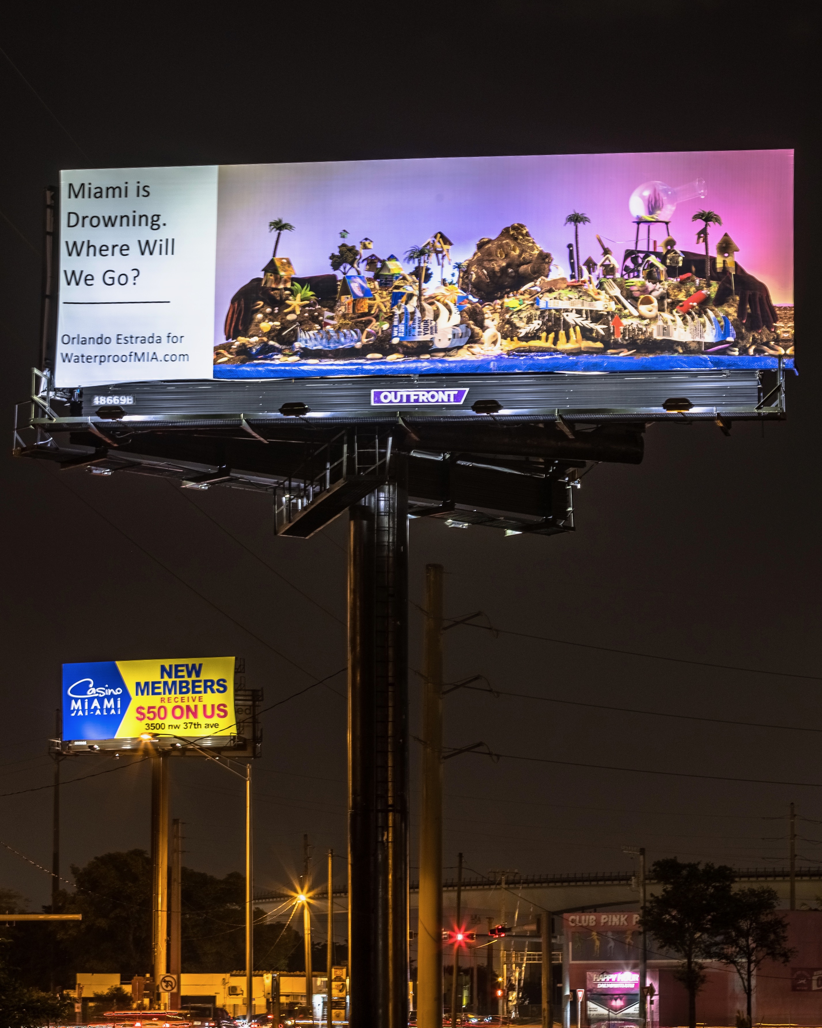 At night, a billboard is displaying an island sunset with a tropical heaven on the left side and a dystopian hell on the right beside a text box that reads in black, “Miami is Drowning. Where Will We Go? Orlando Estrada for WaterproofMIA.com”.