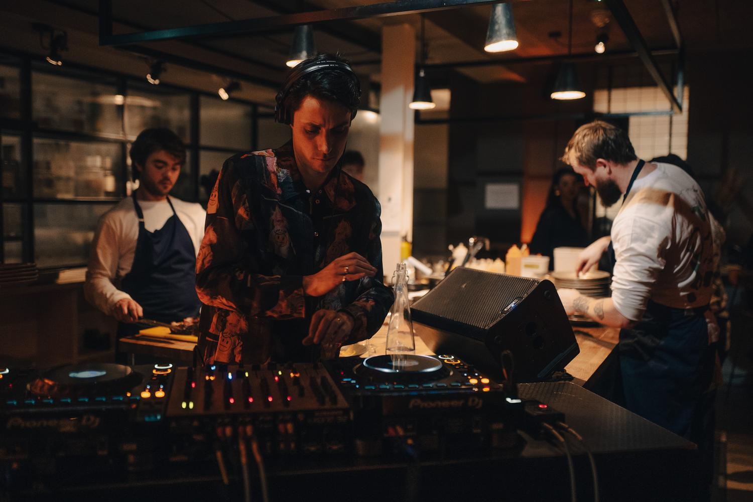 DJ performing beside the open kitchen while chefs plate dishes during a Paired evening at Carousel London.