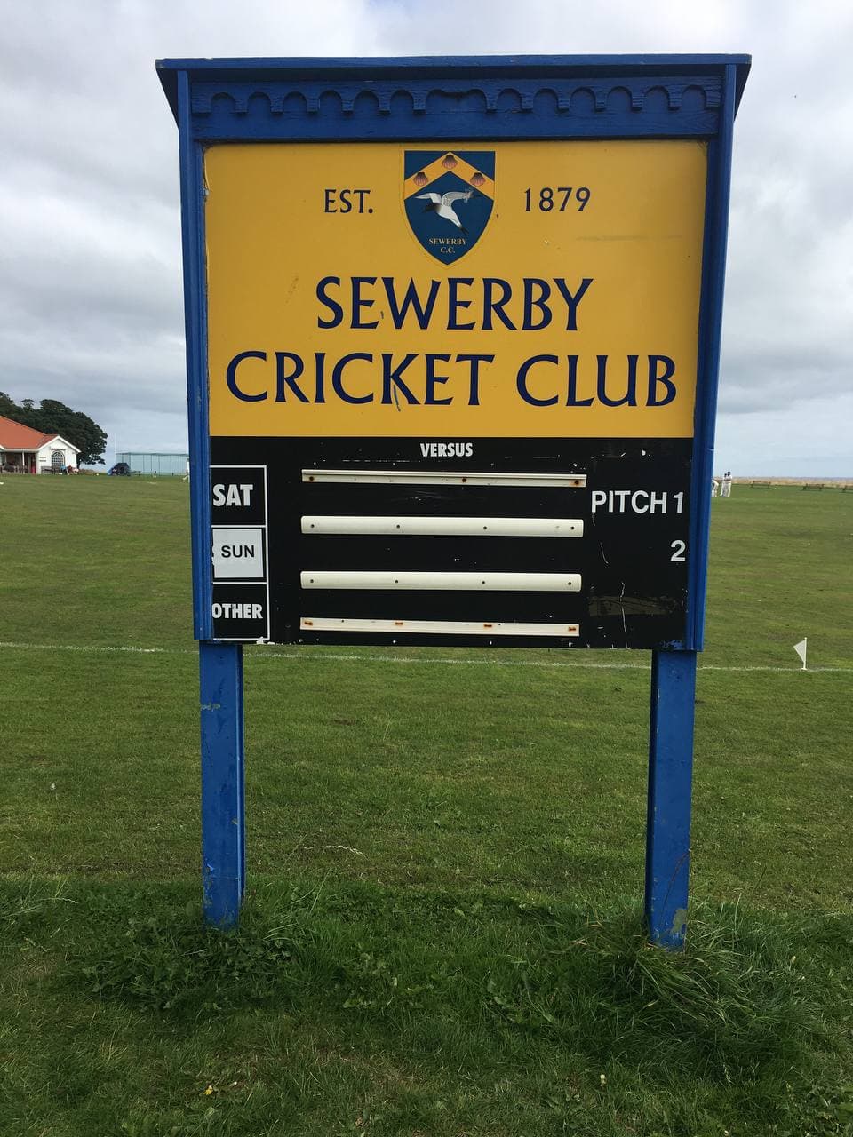 A yellow sign for Sewerby Cricket Club, established 1879 and displaying a coat of arms and listing for Pitch 1.