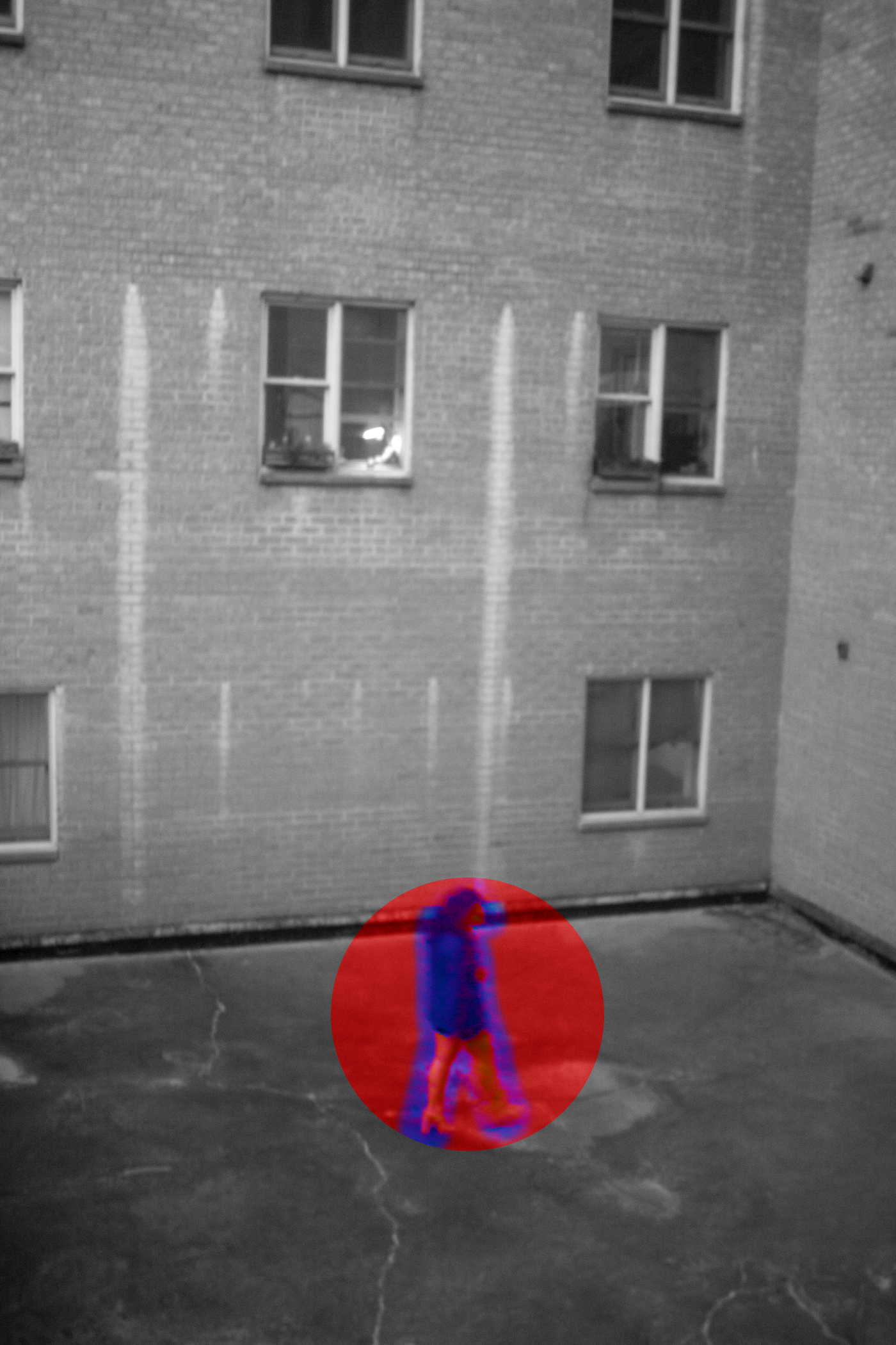 Black-and-white cinematic image of a woman walking through a courtyard, captured from above at a high angle. The eerie, almost surveillance-like composition highlights her figure in blue, encircled in red, creating an infrared effect. Part of the Ambiguous Loss photo book.