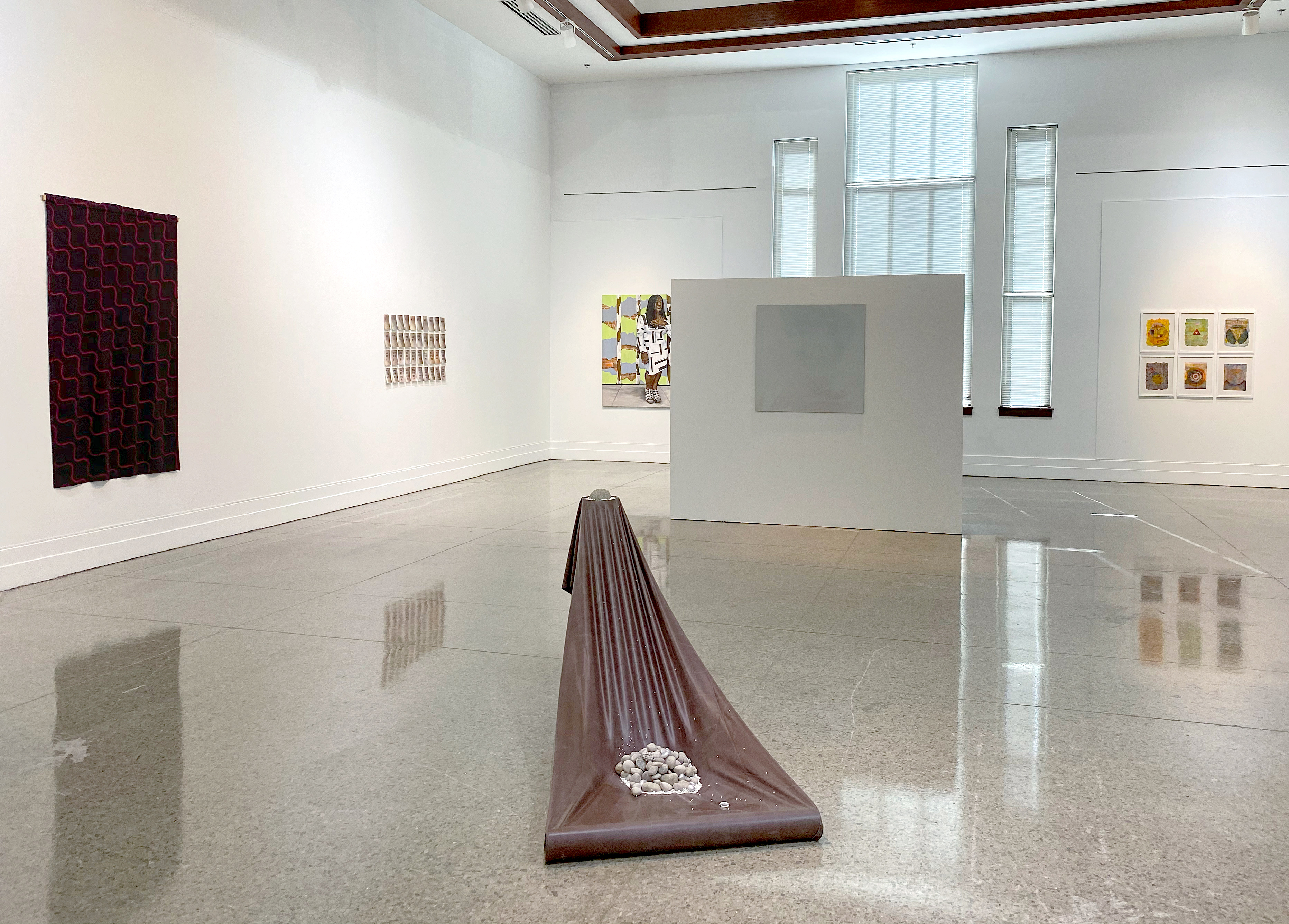 3/4 view of a gallery space, with white walls, a three bay window on the back wall with a shorter, temporary wall in front of it. Art lines the walls and closest to view is a floor sculpture involving stretched brown tarp, held at a 45 degree angle and anchored on either end with rocks and stones.