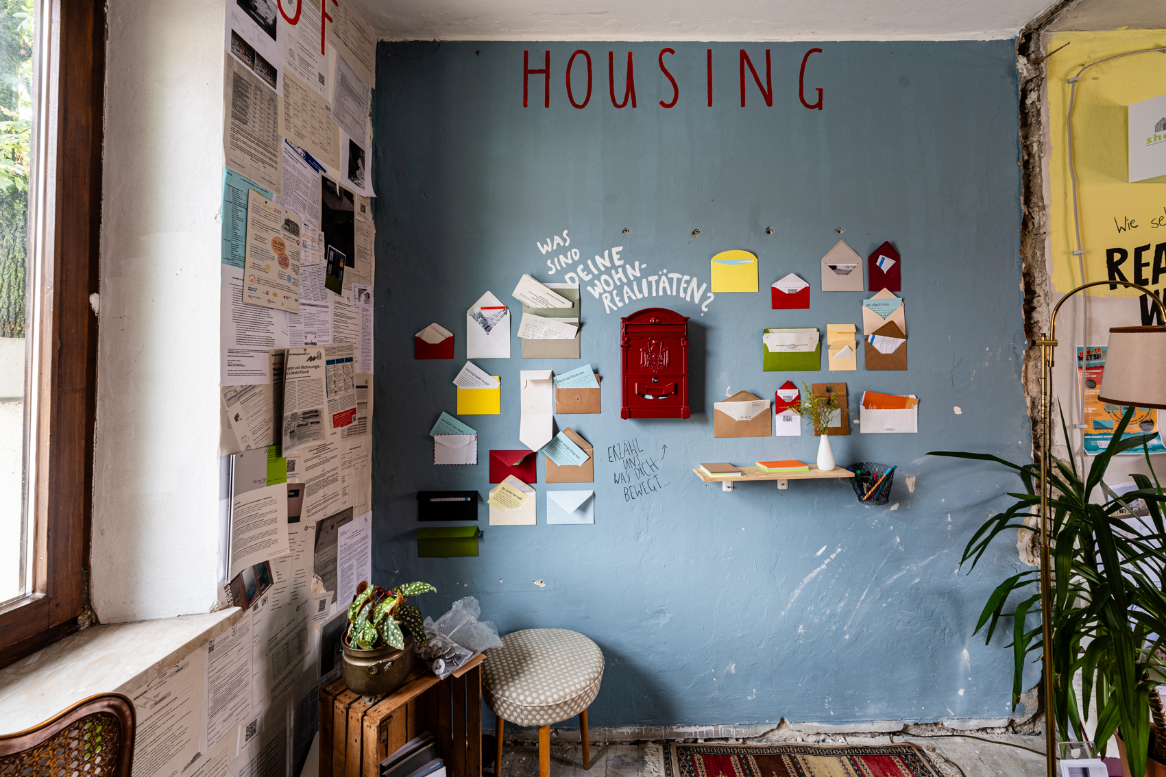 Bild 1 zu Realities of Housing: An einer blauen Wand ist mittig ein roter Briefkasten befestigt. Dar&uuml;ber steht mit wei&szlig;en Buchstaben: "Was sind Deine Wohn-Realit&auml;ten?" An der Wand h&auml;ngen verschiedenfarbige Briefumschl&auml;ge.