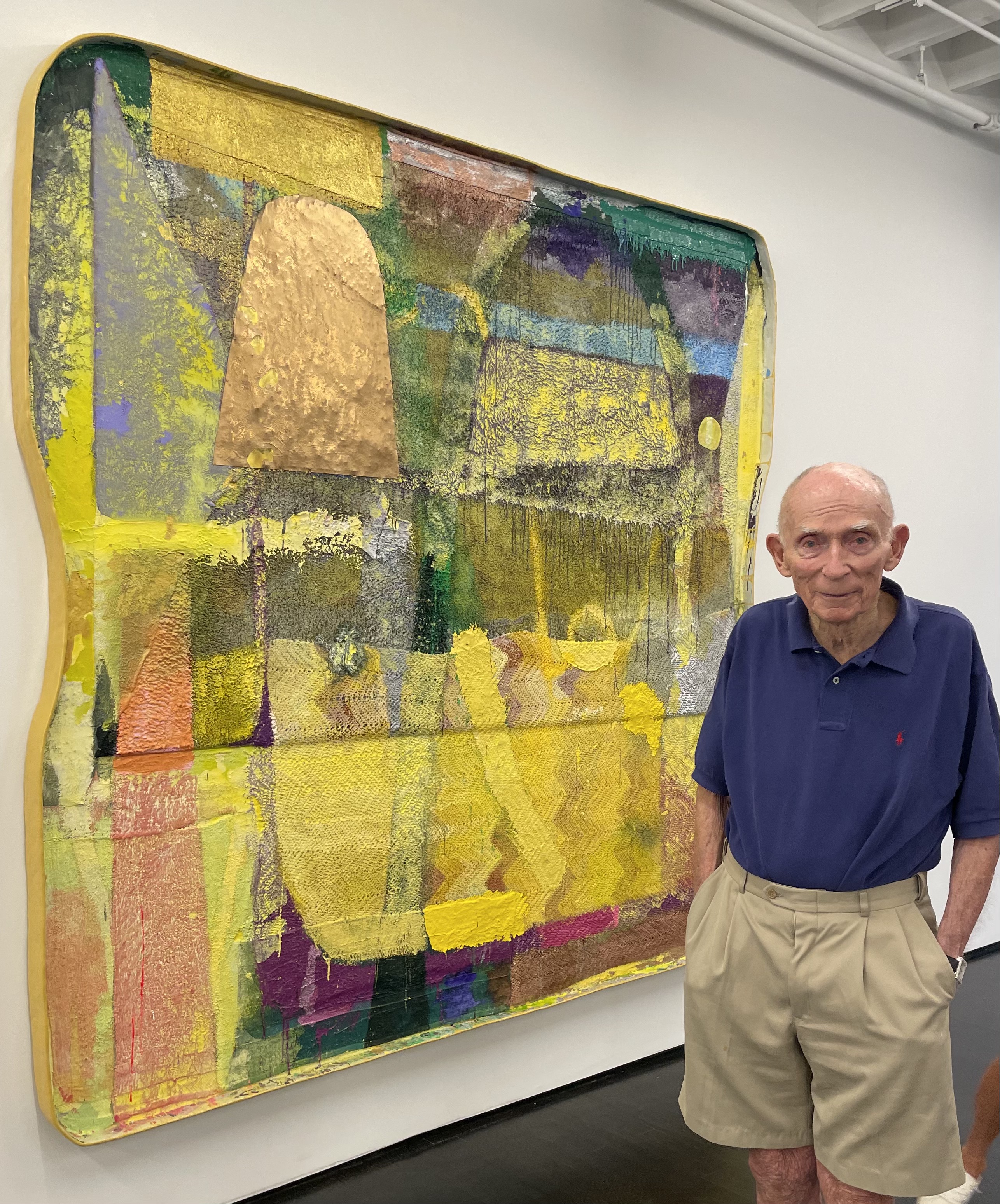 Al Shands, a senior man wearing khaki shorts and a dark navy polo is standing in front of a large abstract rectangular painting with rounded corners, heavily textured, with colors of peach, yellow, grey, blue and purple.