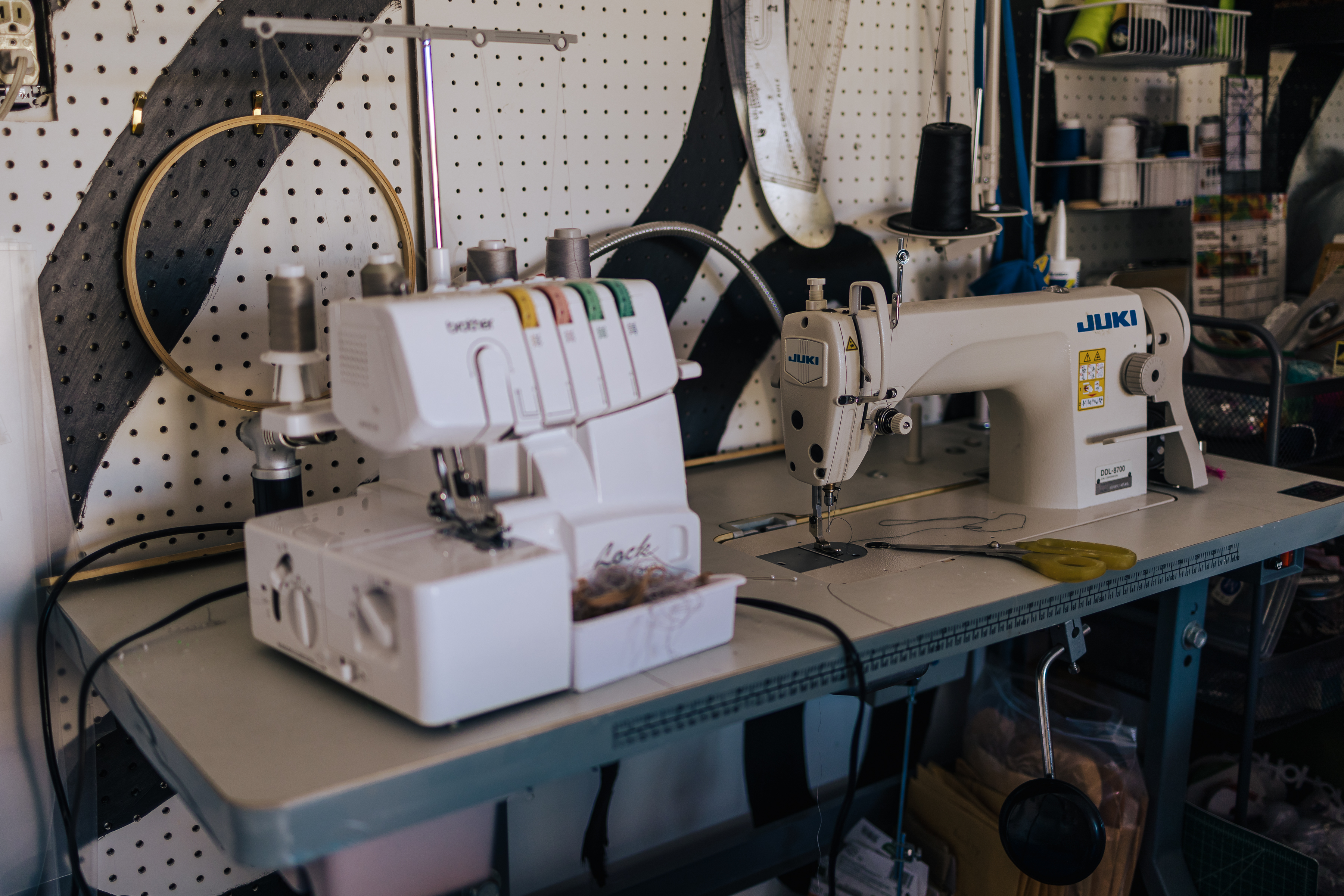 Photo by Mario Ramirez of the sewing table, pictured is an industrial sewing machine, overlock machine, and behind the table is a tool wall with embroidery  hoops, rulers, and thread