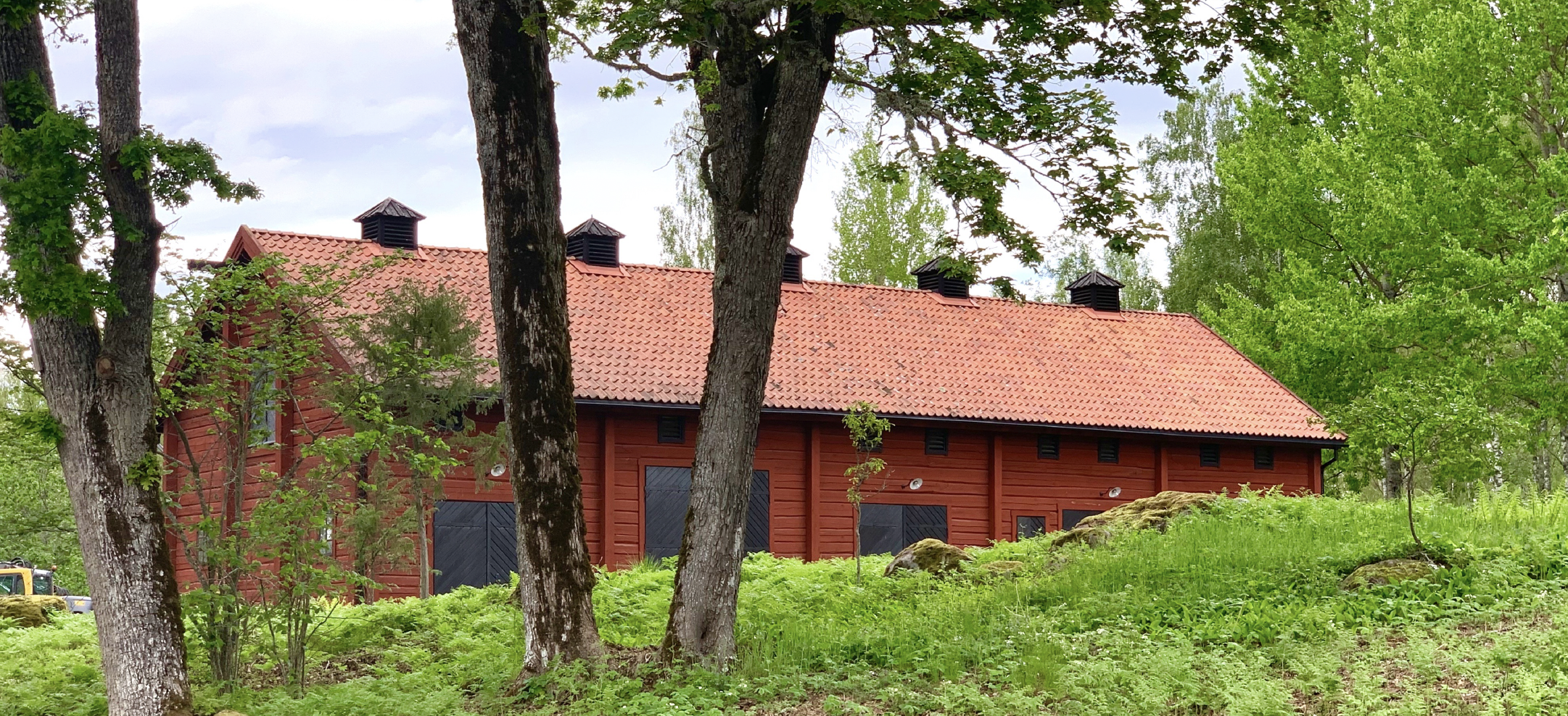 Stallet är diskret infogat i naturen i lummig grönska. Falurött liggtimmer med knutar, enkupigt rött lertegel hör till byggtraditionen även här.