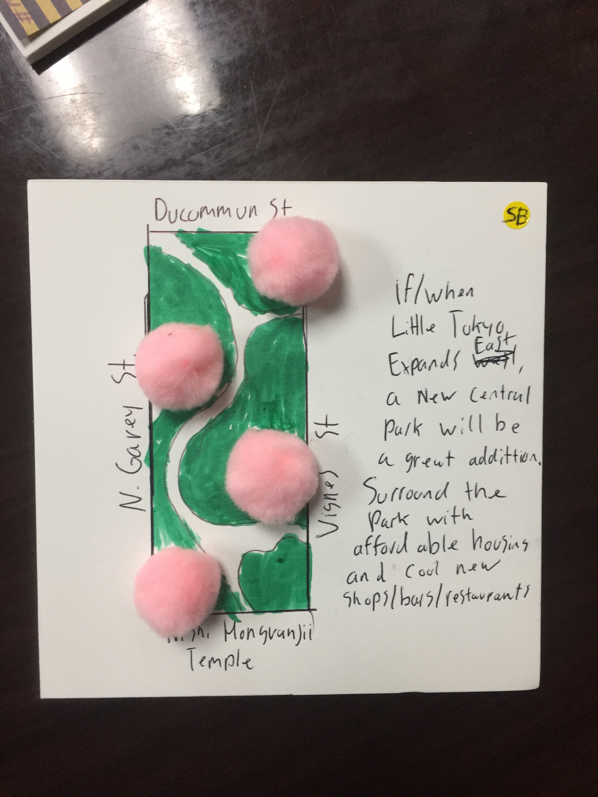 photo shows a small 6x6 square with a hand drawn map of green space and pink pom poms, made by a community member at a design workshop. They included a written description of their idea: If/when Little Tokyo expands east, a new central park will be a great addition. Surround the park with affordable housing and cool new shops/bars/restaurants.