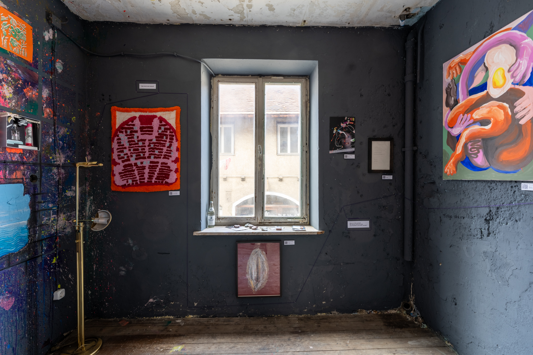 Bild 1 zu Mehr Raum f&uuml;r Wut: Ein quadratischer Raum in grau mit mehreren Gem&auml;lden an den W&auml;nden. Der Raum hat ein Fenster an der gegen&uuml;berliegenden Seite der T&uuml;r. Links vom Fenster h&auml;ngt ein Kunstwerk aus F&auml;den, in Orange, Rosa und Rott&ouml;nen. 