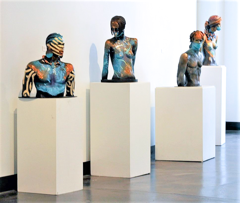 Four figurative sculptures, all from the waist up, line a wall in a gallery and each sit on top of white podiums of various heights. The figures are modeled realistically, but theirs surfaces have expressive splashes of colors and geometric patterns all over them.