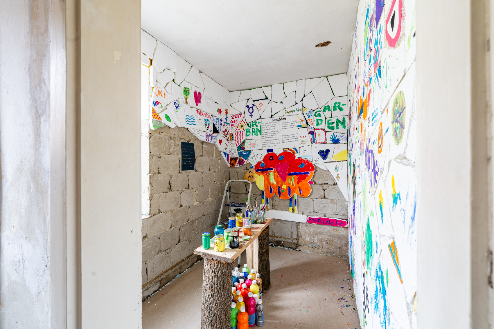 Bild 2: Zwischen schwarz und wei&szlig;: Der Raum wurde mit einzelnen Styroporst&uuml;cken verkleidet, die von Besuchenden bemalt wurden. Davon ist die rechte Wand komplett bedeckt, der Rest nur teilweise, sodass die Steinwand des Hauses zu sehen ist. In der Mitte des Raumes steht ein Holztisch auf dem viele bunte Farben stehen.