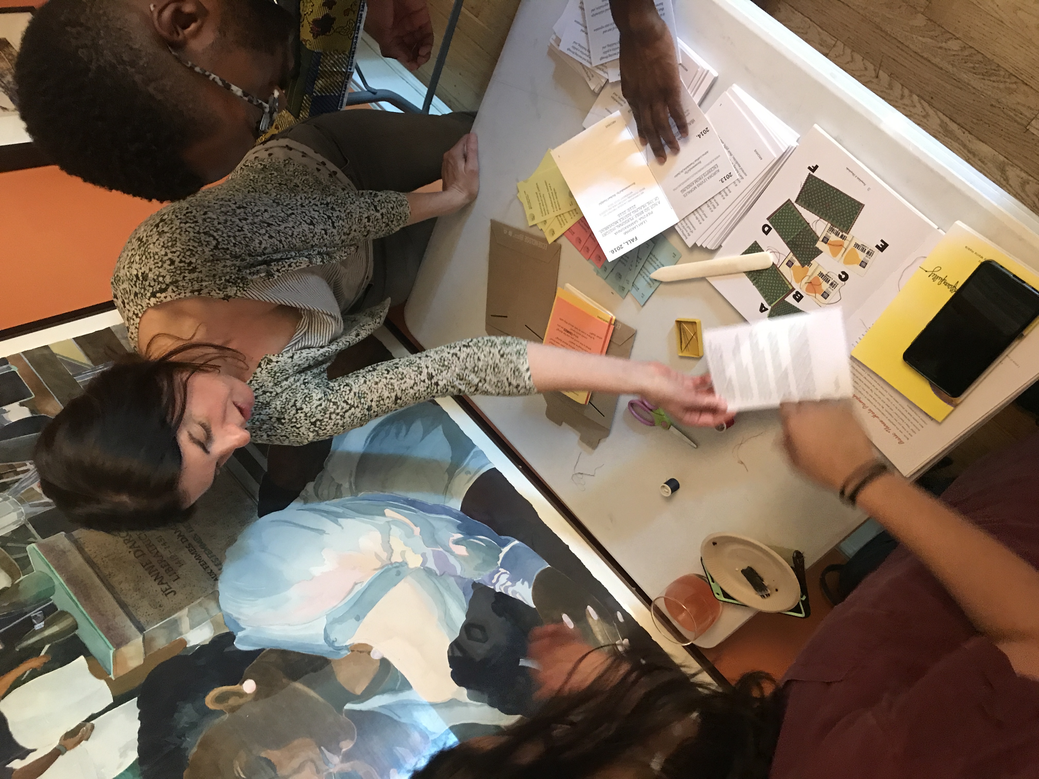  Photo of 3 people folding books in front of a colorful painting--a young Black man, a middle-aged white woman, and somebody whose long hair is visible. Photo of butcher block paper with words written in blue marker: &ldquo;What does healing justice mean to you?&rdquo; Some answers are difficult to make out. Readable ones say, &ldquo;Restoration of culture and & authentic expression. Emotional embodiment + intimacy.&rdquo; &ldquo;Love for one another no matter what. Give space. Exist beyond labels.