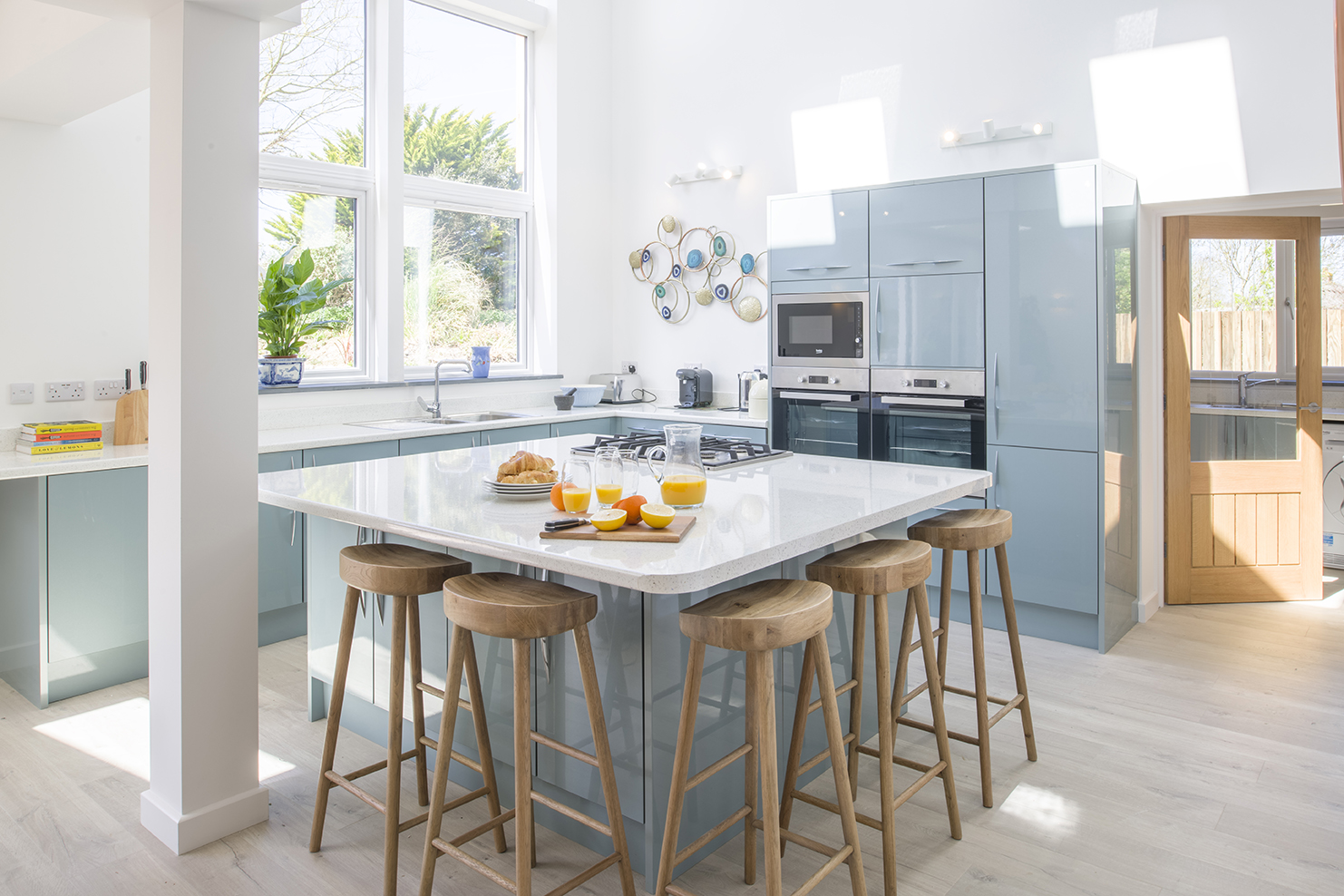 Light blue kitchen units and breakfast bar with white surfaces. Wooden stools. High ceilings creating a bright, airy space.